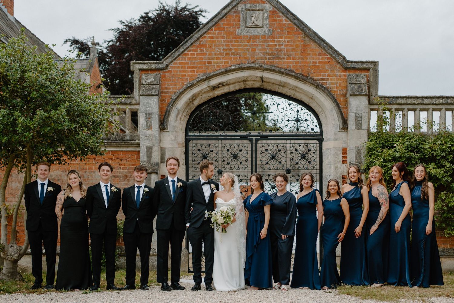 Wedding party group photo with bride, groom, groomsmen in black tuxedos, and bridesmaids in navy gowns before ornate iron gates.