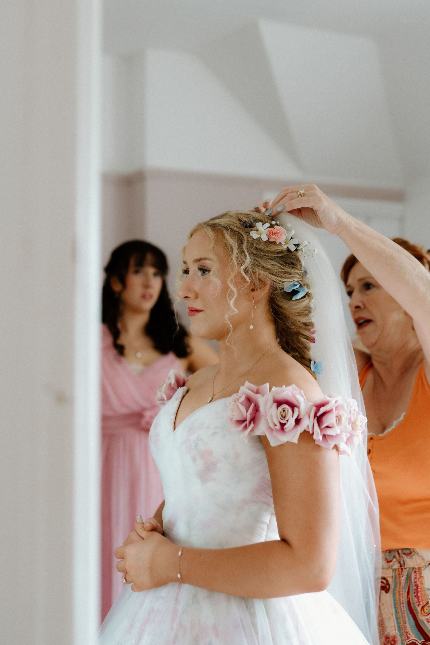 Mother helping bride with veil while bridesmaid watches in background during wedding preparations.