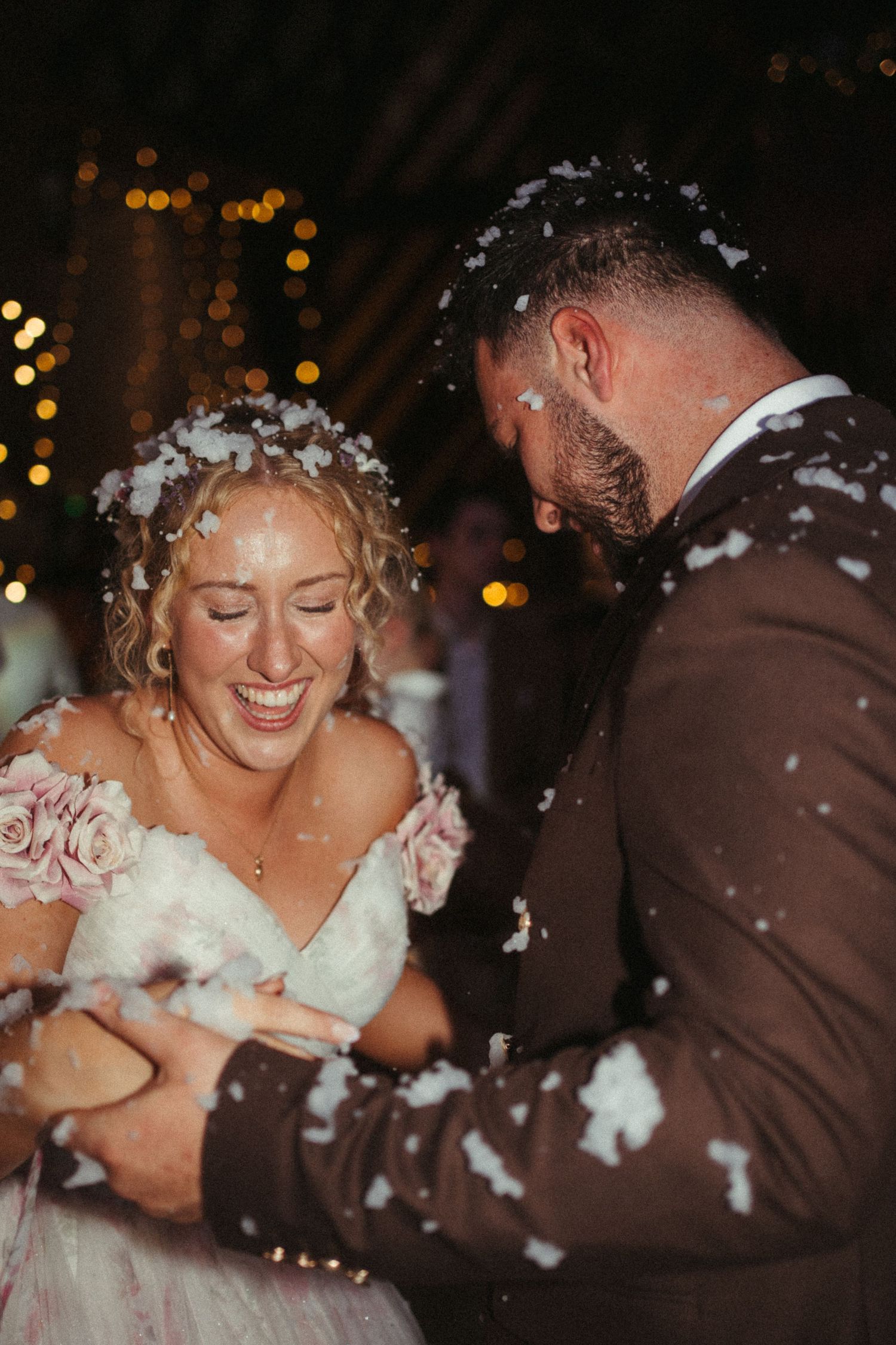 Joyful bride and groom laugh together as confetti falls around them during wedding celebration.