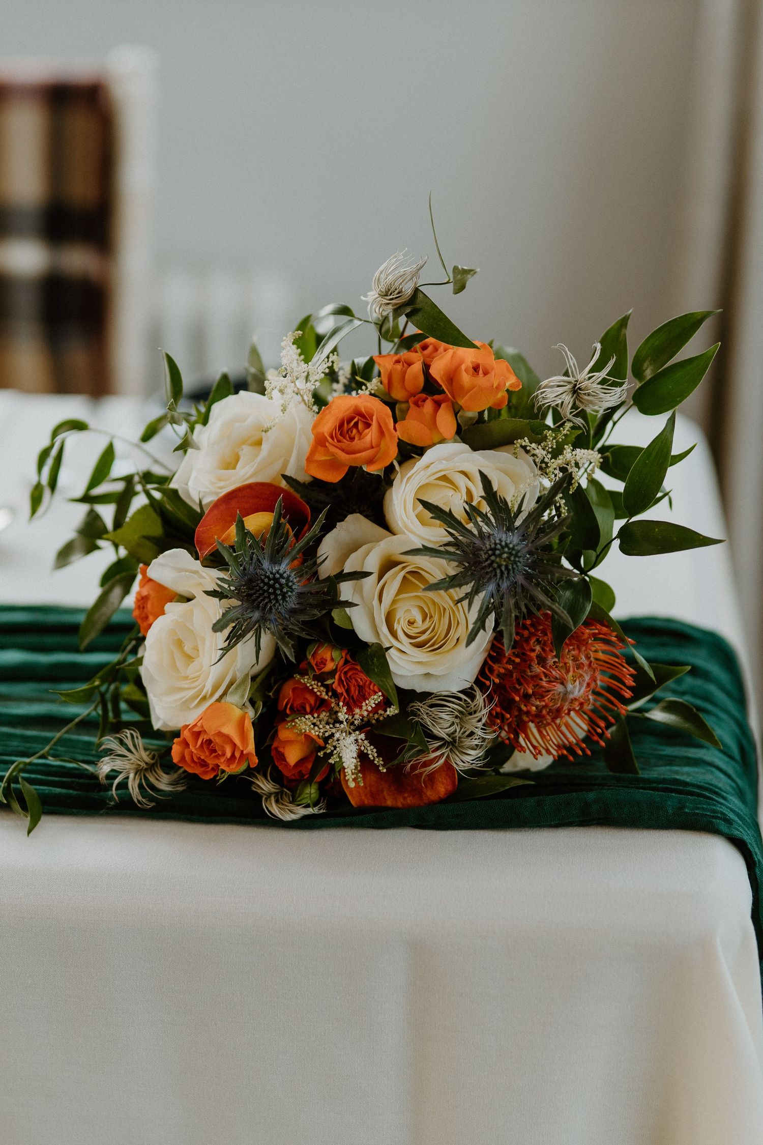 Bridal bouquet with orange and white roses, thistles, and greenery on green velvet table runner.