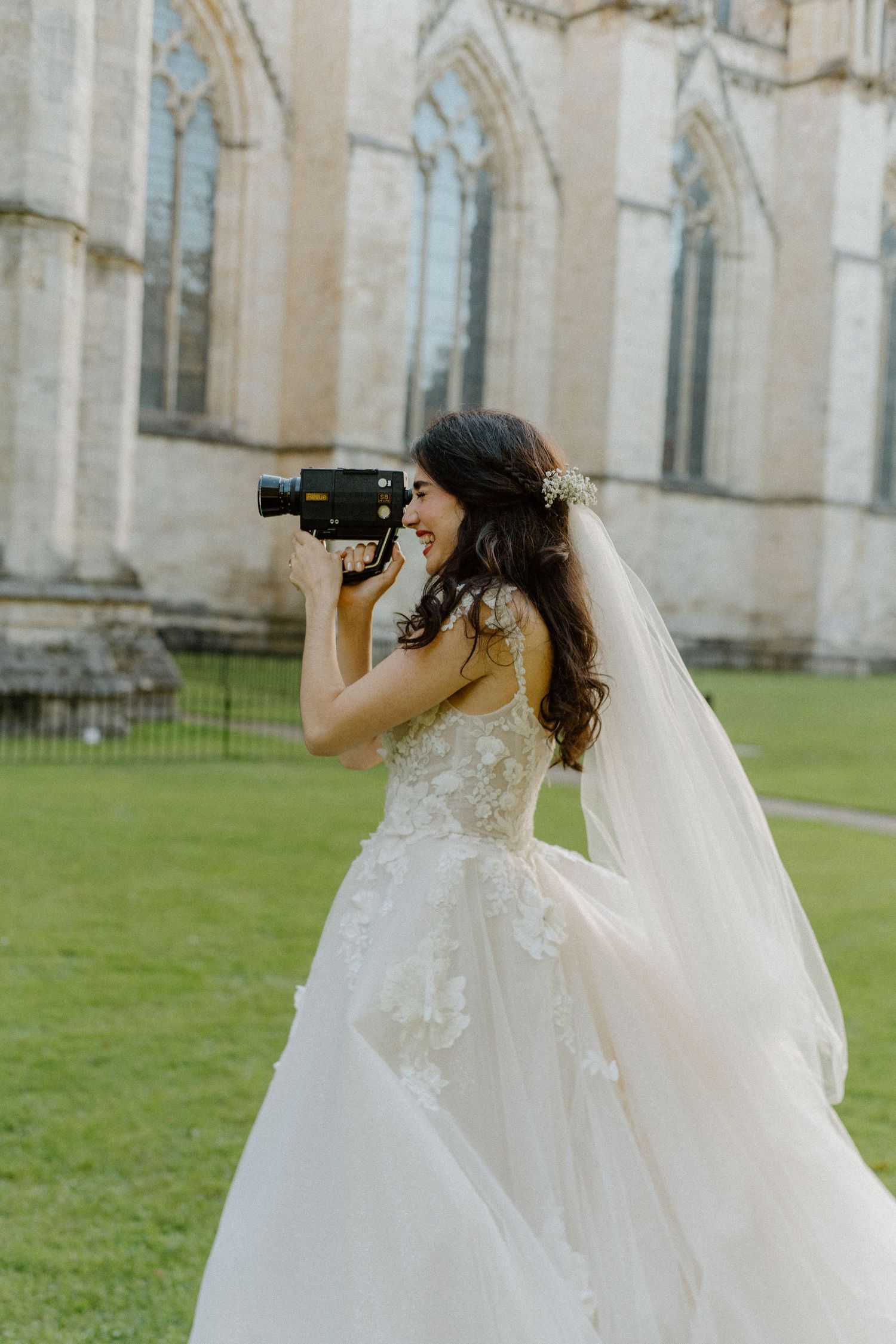 Smiling bride in lace wedding gown and veil filming with vintage camera outside historic stone cathedral.