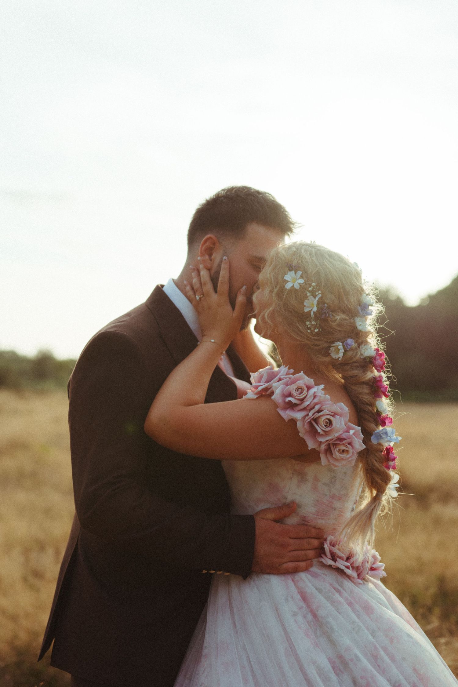 A romantic wedding couple sharing a tender kiss at sunset in a field, the bride wearing a floral crown and tulle dress.