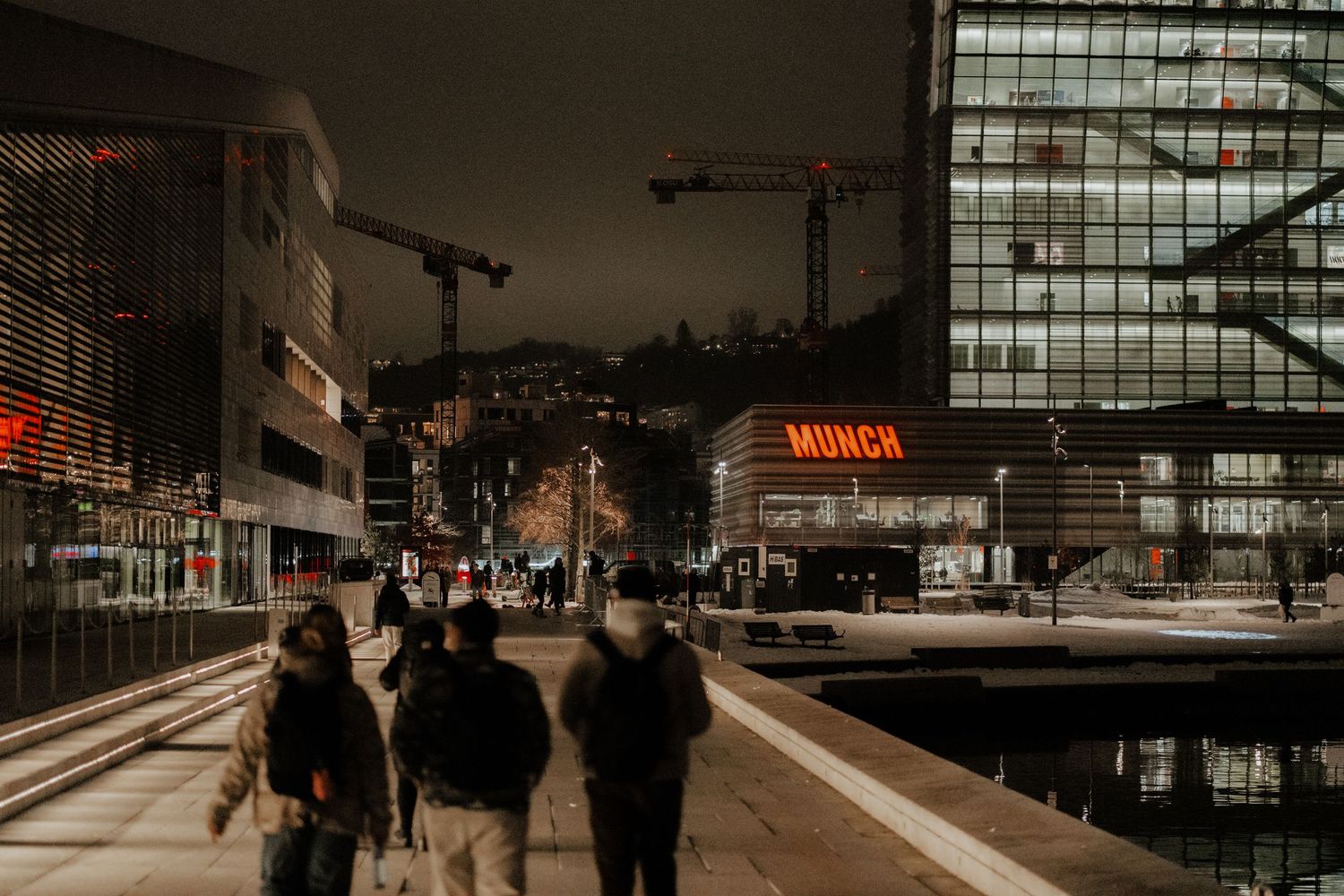 People walking at night along an urban street with illuminated buildings and construction cranes in Amsterdam.