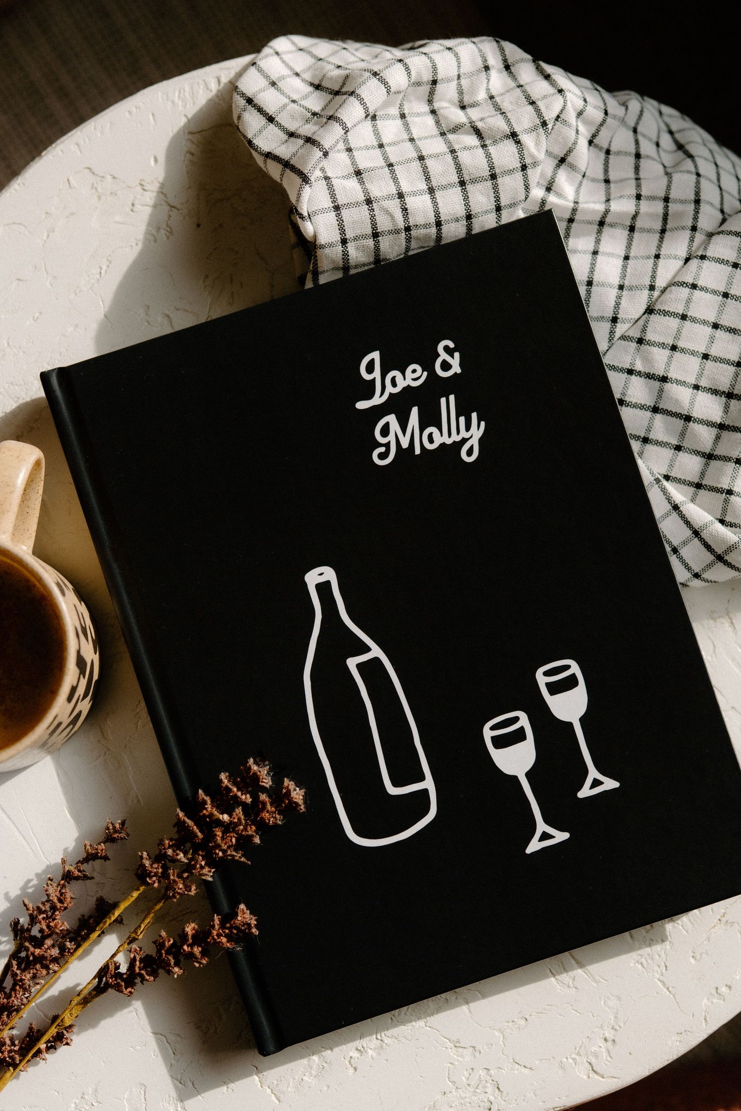 Black book cover with wine bottle and glasses illustration, names Joe & Molly on display with checkered cloth backdrop.