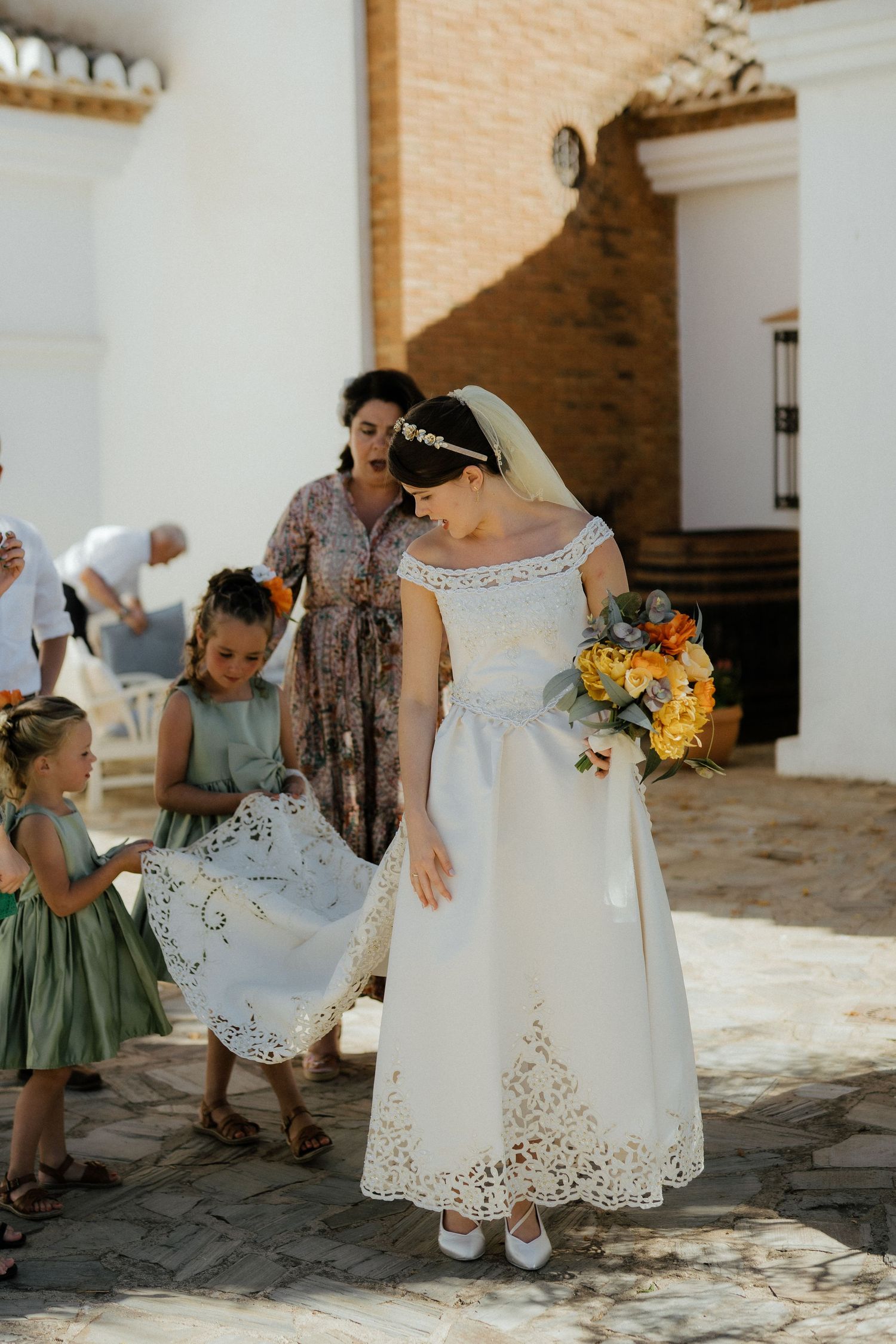 Bride in white lace gown walks with flower girls in sage green dresses outside a whitewashed Spanish venue.