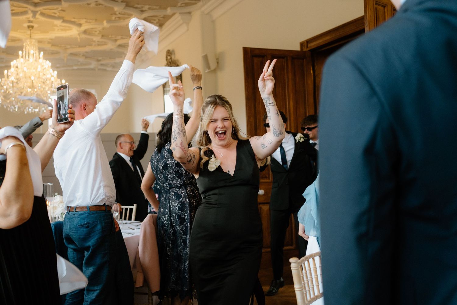 Excited wedding guest in black dress celebrating with raised arms during a lively wedding reception entrance.