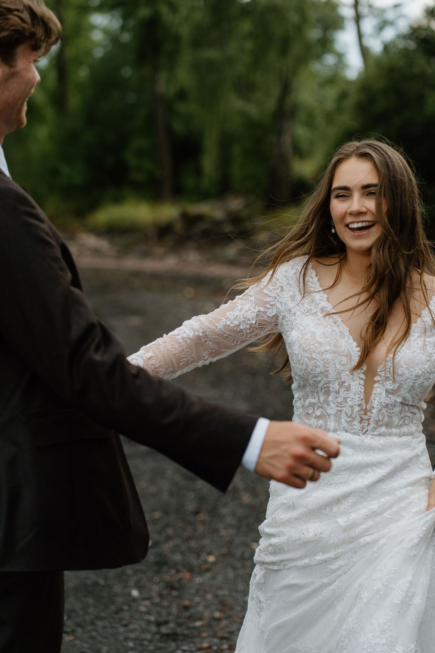 Smiling bride in beaded lace gown holds grooms hand while walking outdoors on wedding day.
