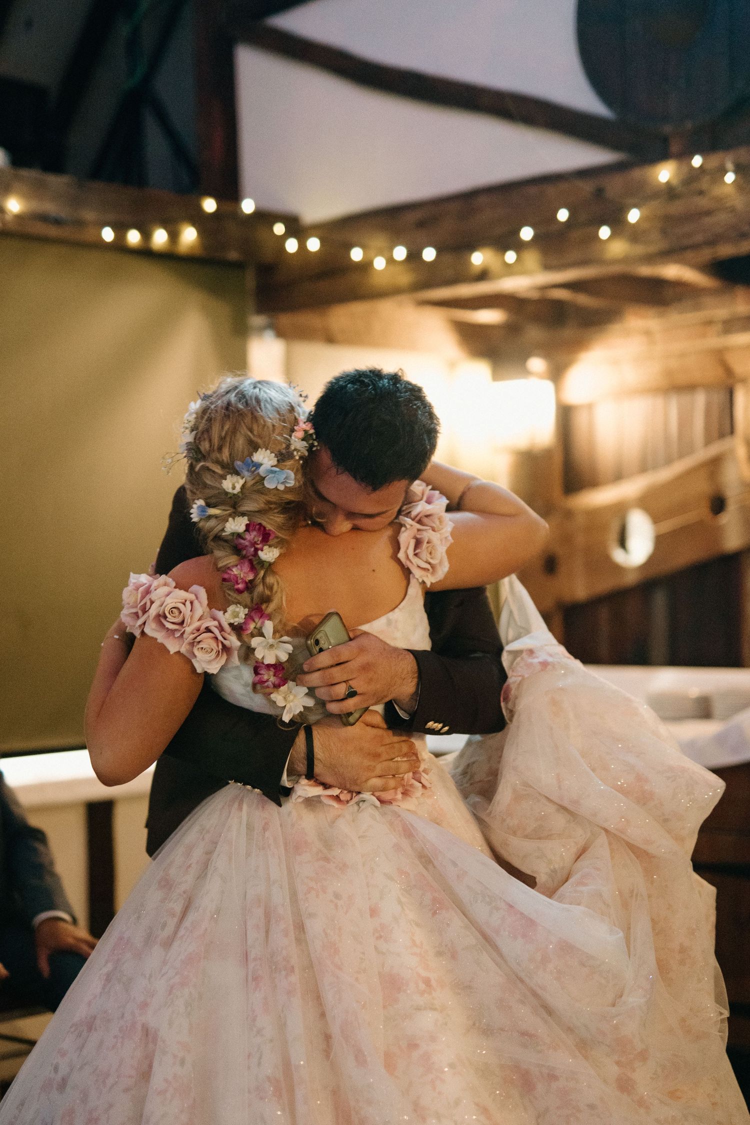 Bride and groom share emotional first dance embrace in warmly lit barn decorated with string lights.