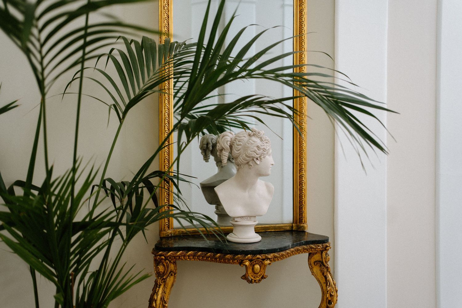 Classical white bust sculpture on a marble console table beside tropical palm leaves in front of a gold-framed mirror.
