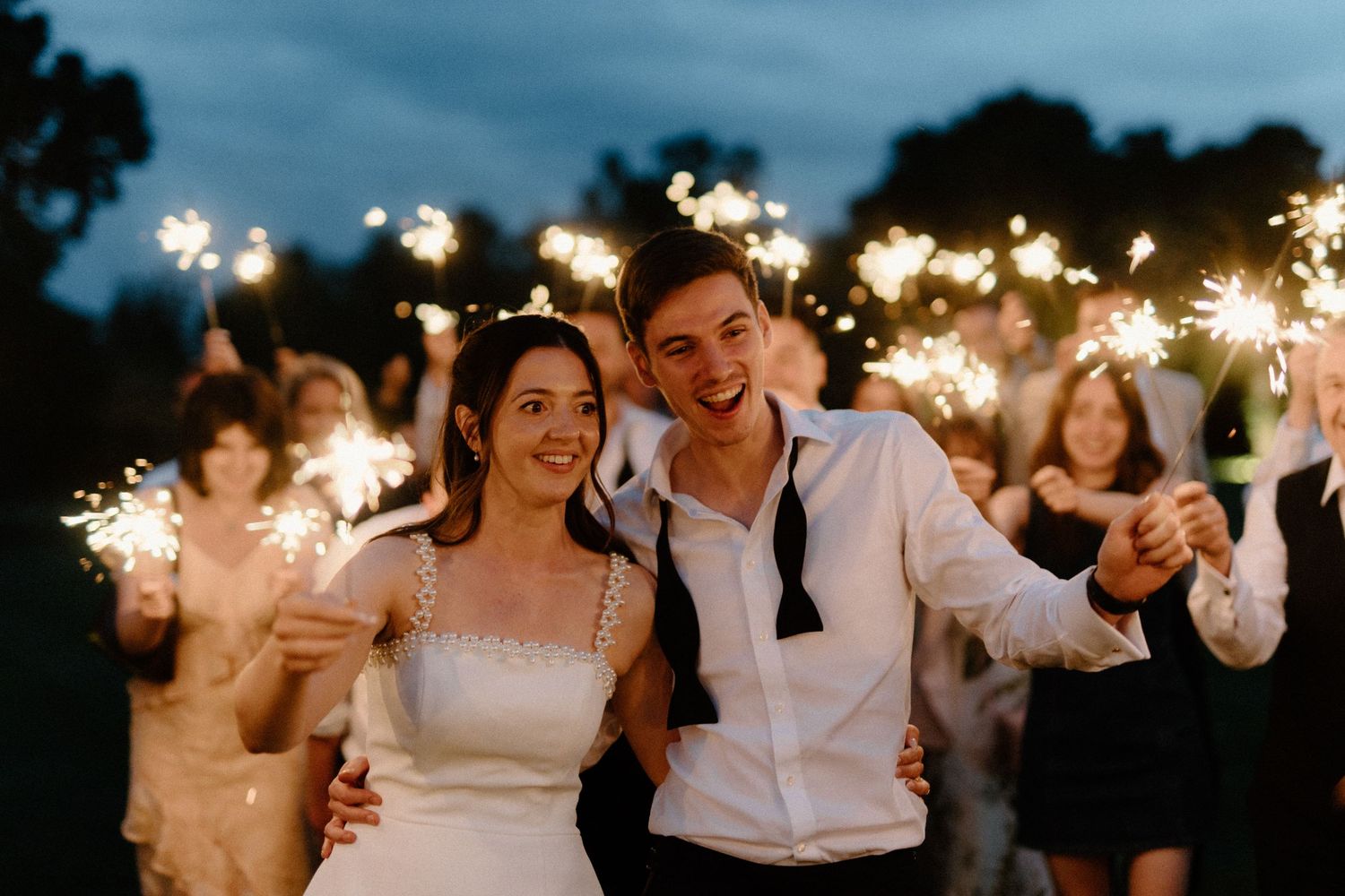 Newlywed couple smiling surrounded by guests holding sparklers during magical outdoor evening send-off.