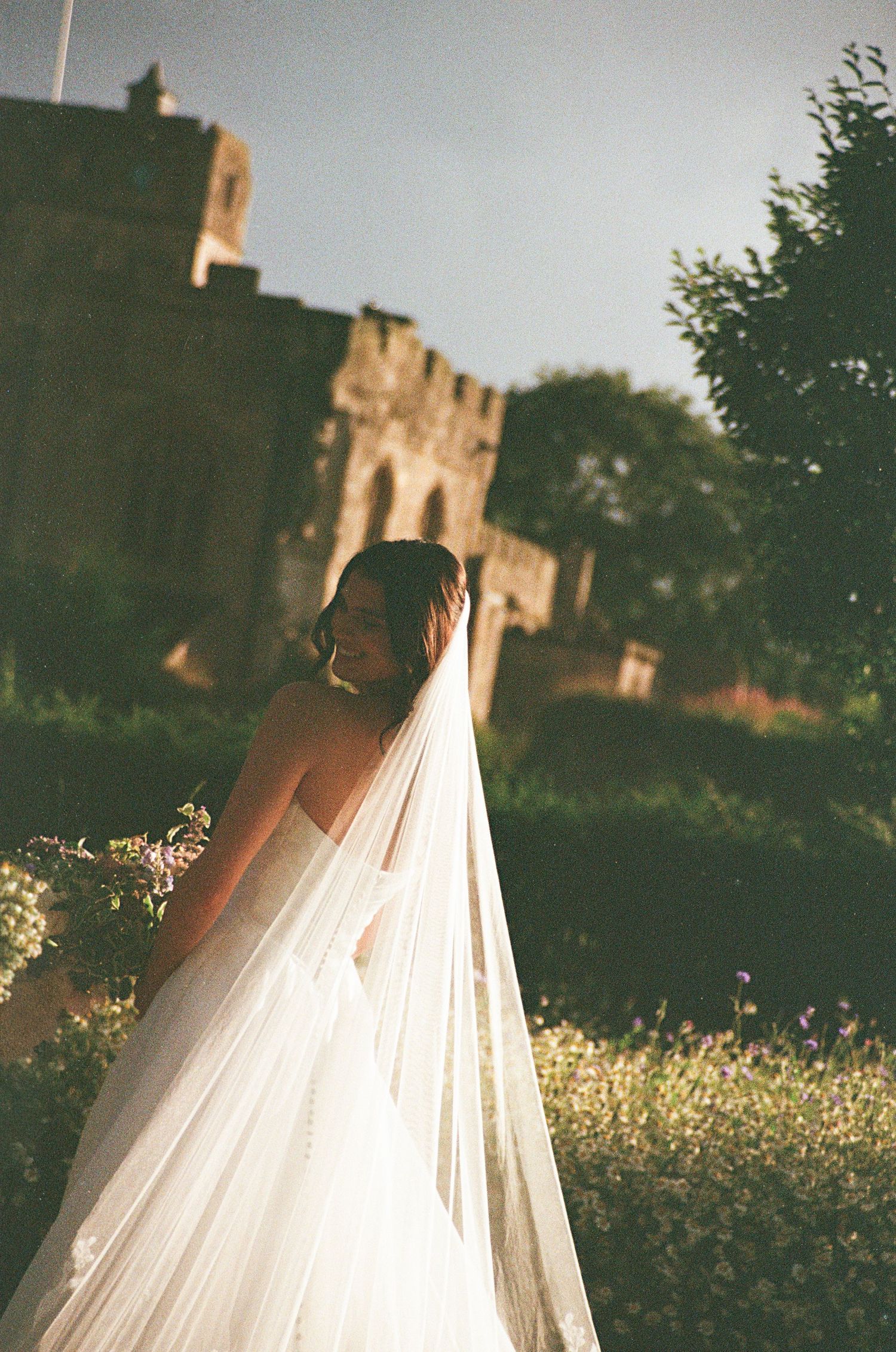 Bride in strapless gown with long veil posing near historic stone castle ruins.