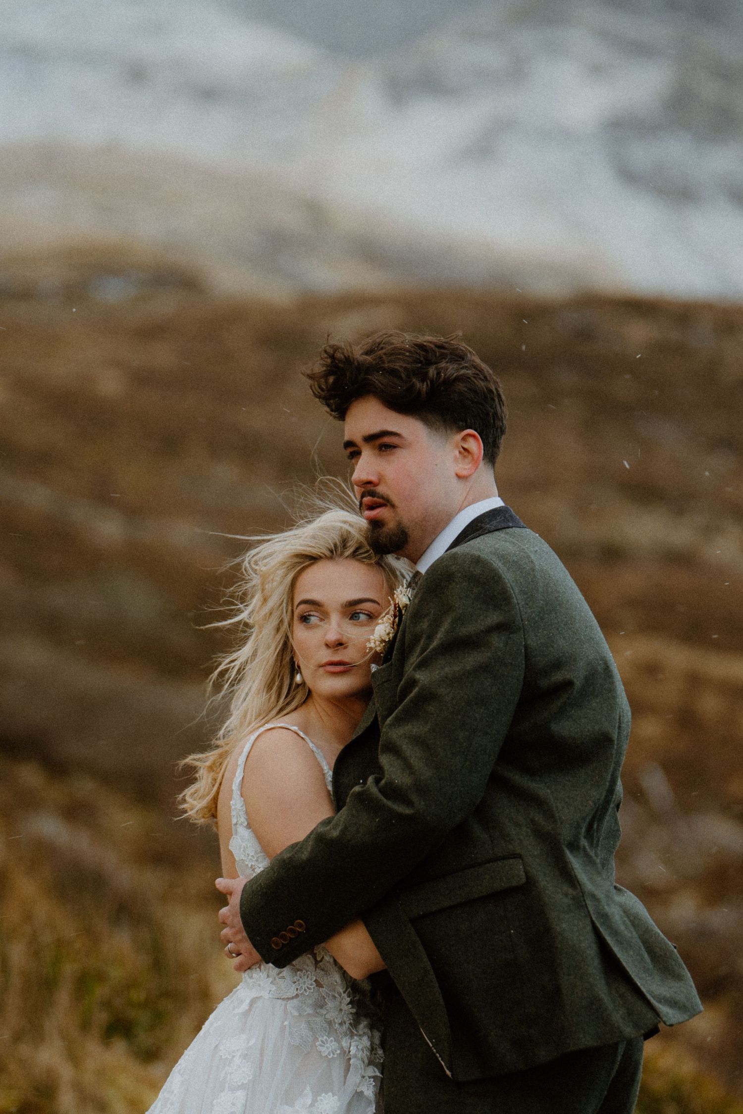 Bride and groom embrace in windy Scottish Highlands, groom wearing green tweed suit outdoors.