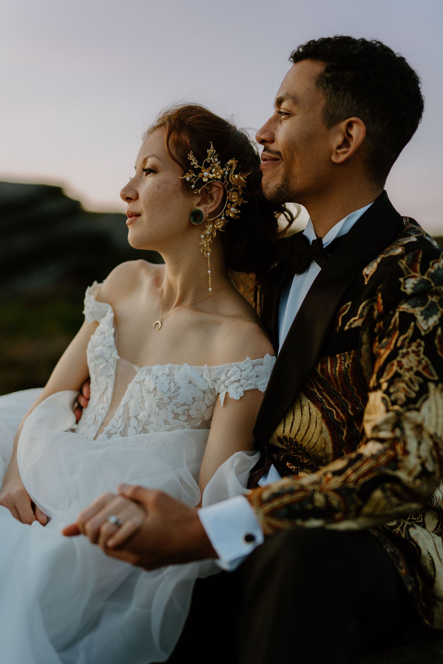 Wedding couple sitting together at sunset, bride with gold hair accessories and groom in decorative gold jacket.