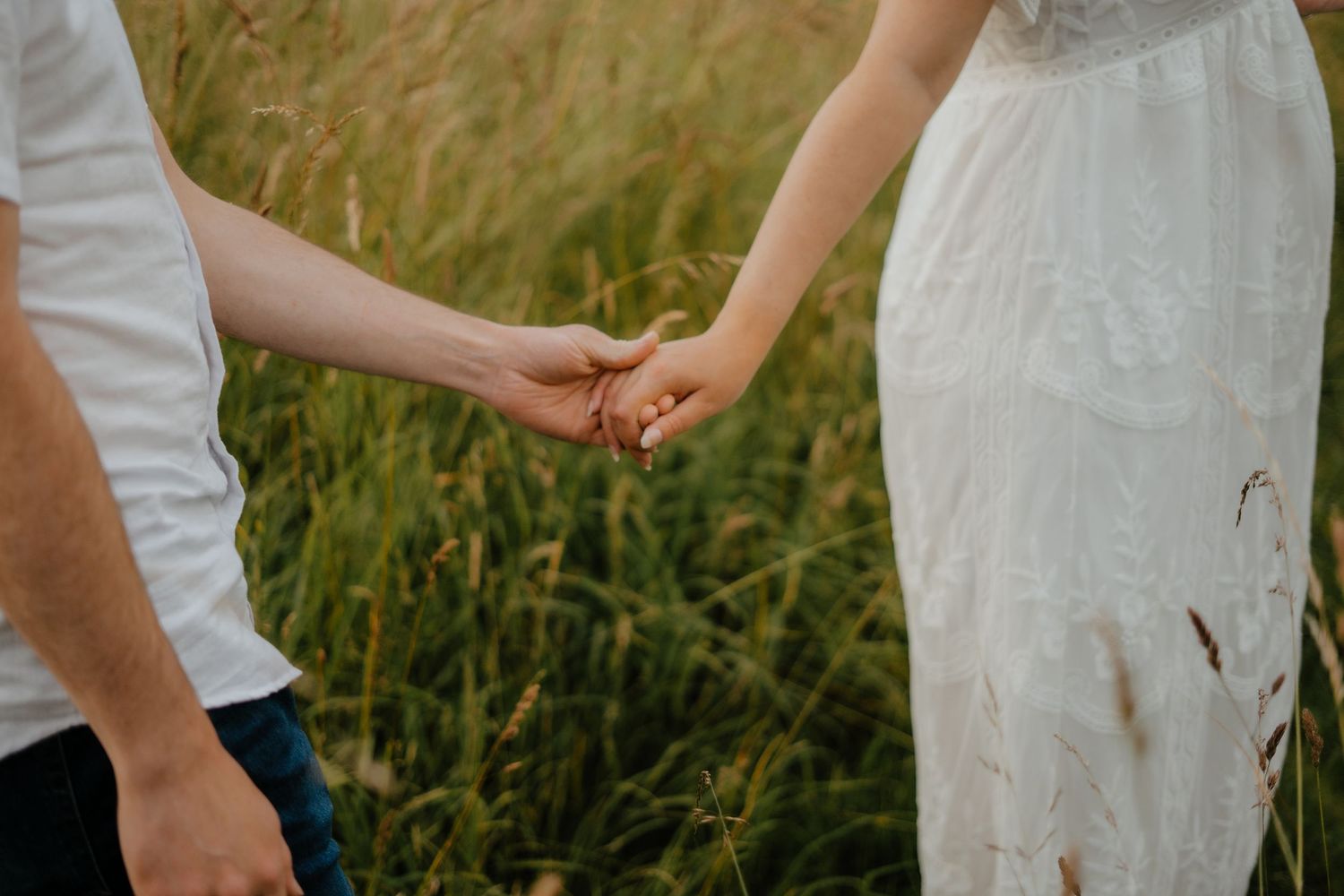 A couple holds hands while walking through tall green grass in a field.
