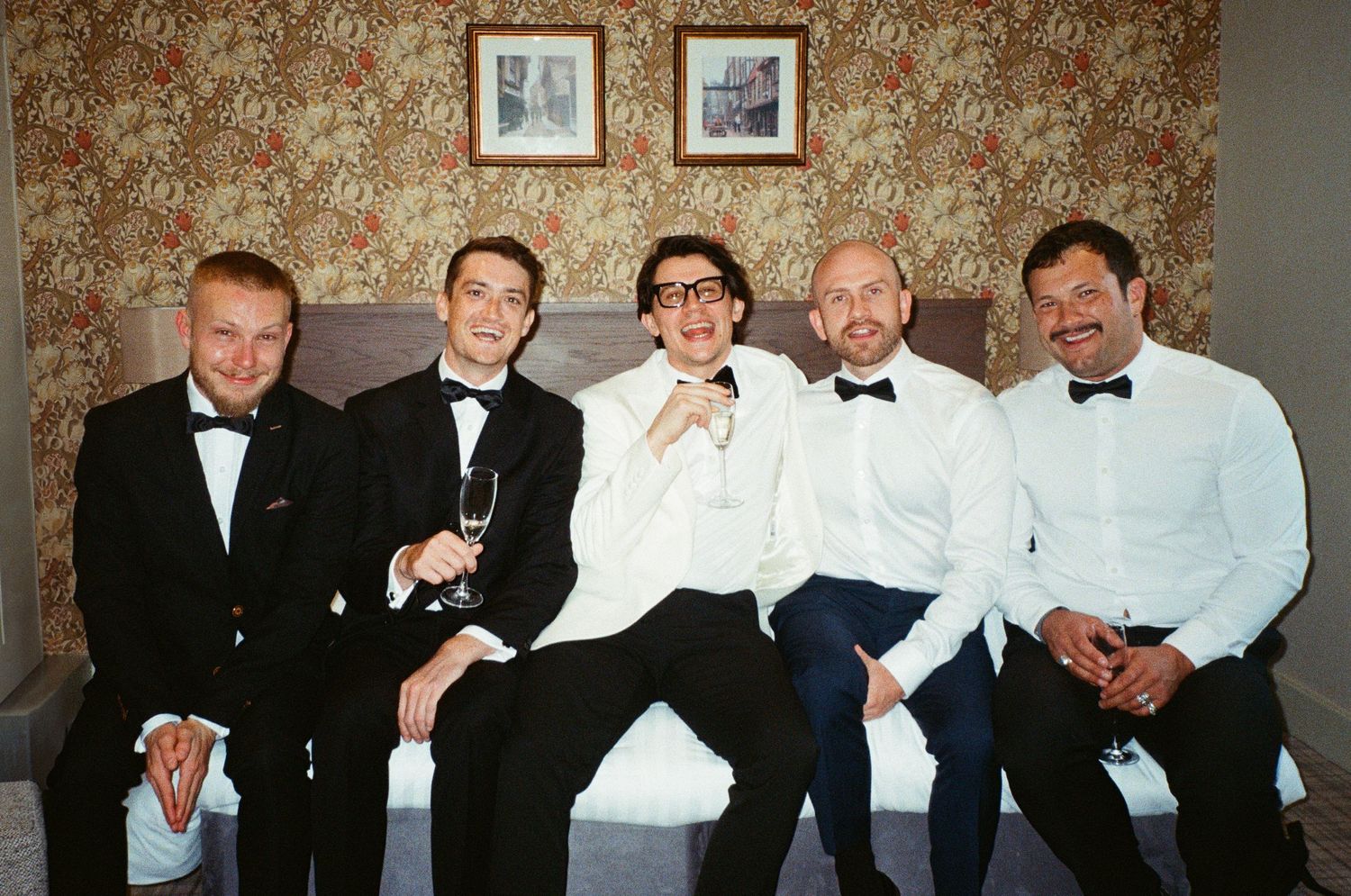 Five groomsmen in tuxedos sitting on a bed smiling, one holding a champagne glass.