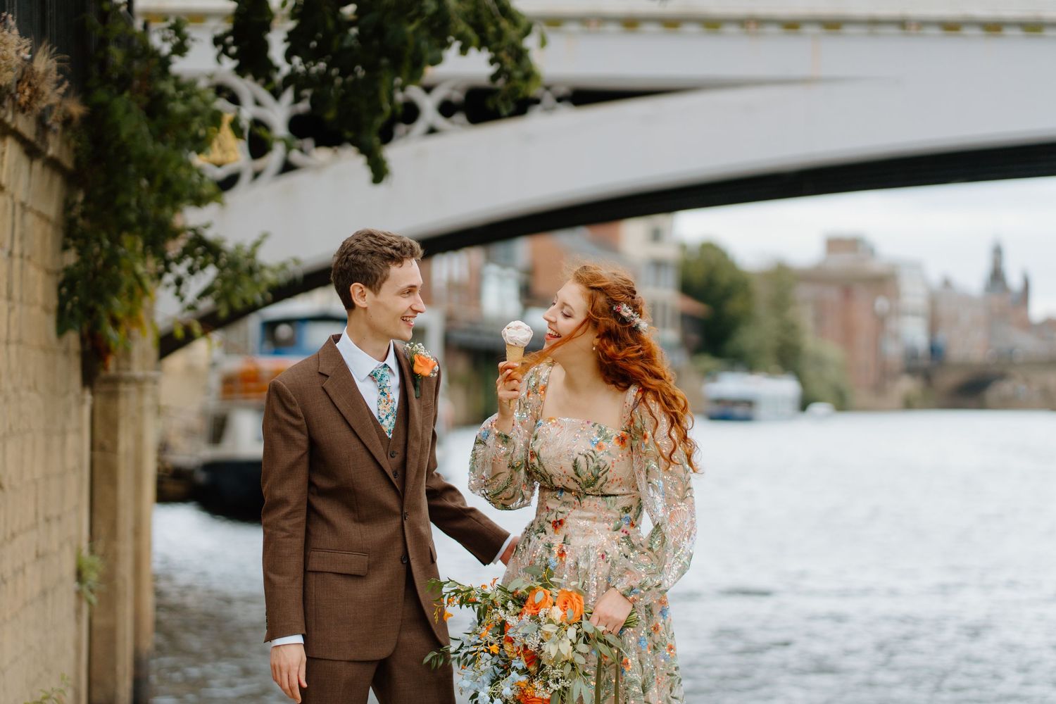 Bride in floral dress and groom in brown suit laugh together by riverside bridge on wedding day.