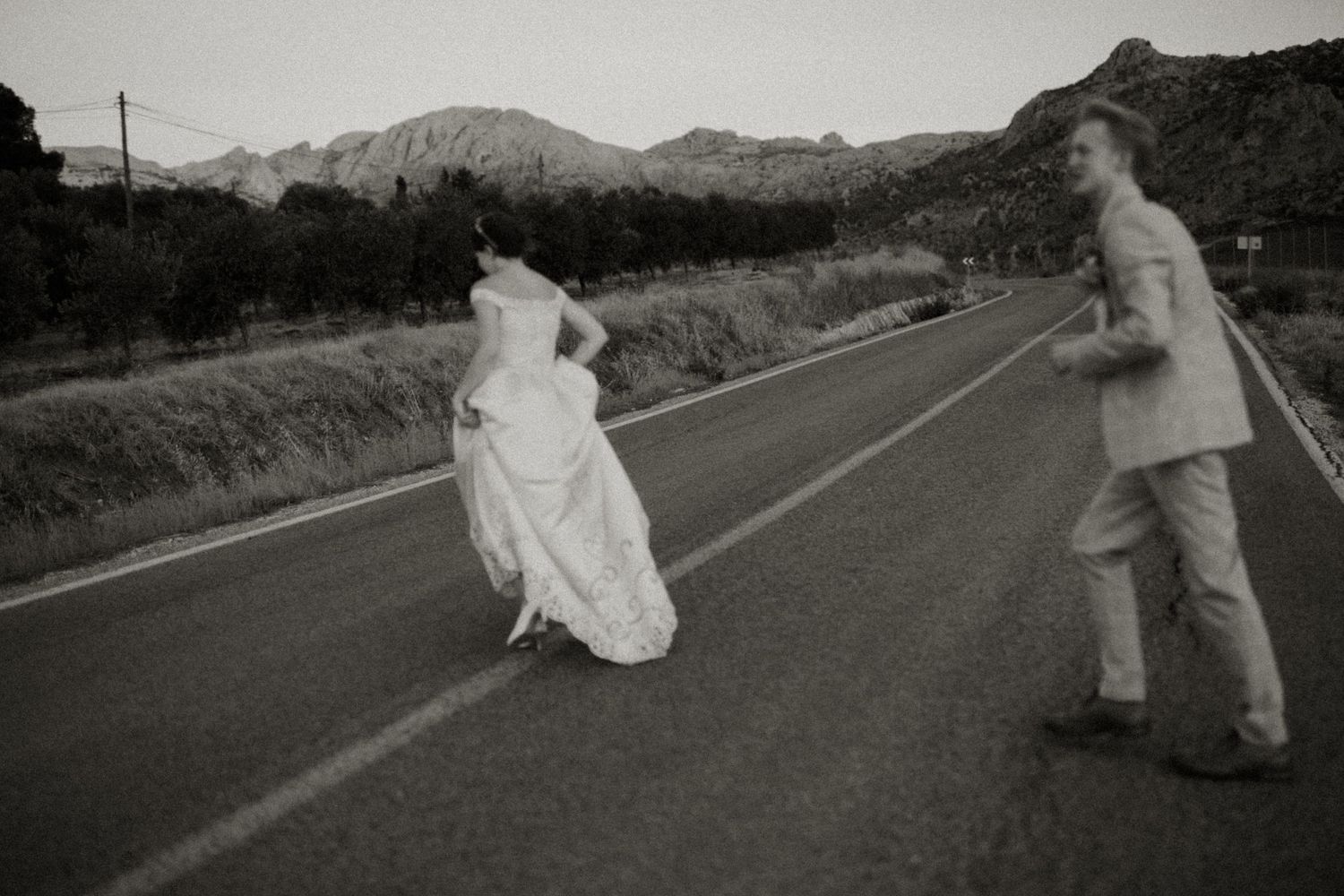 Black and white photo of bride and groom strolling on empty road with dramatic mountain backdrop.