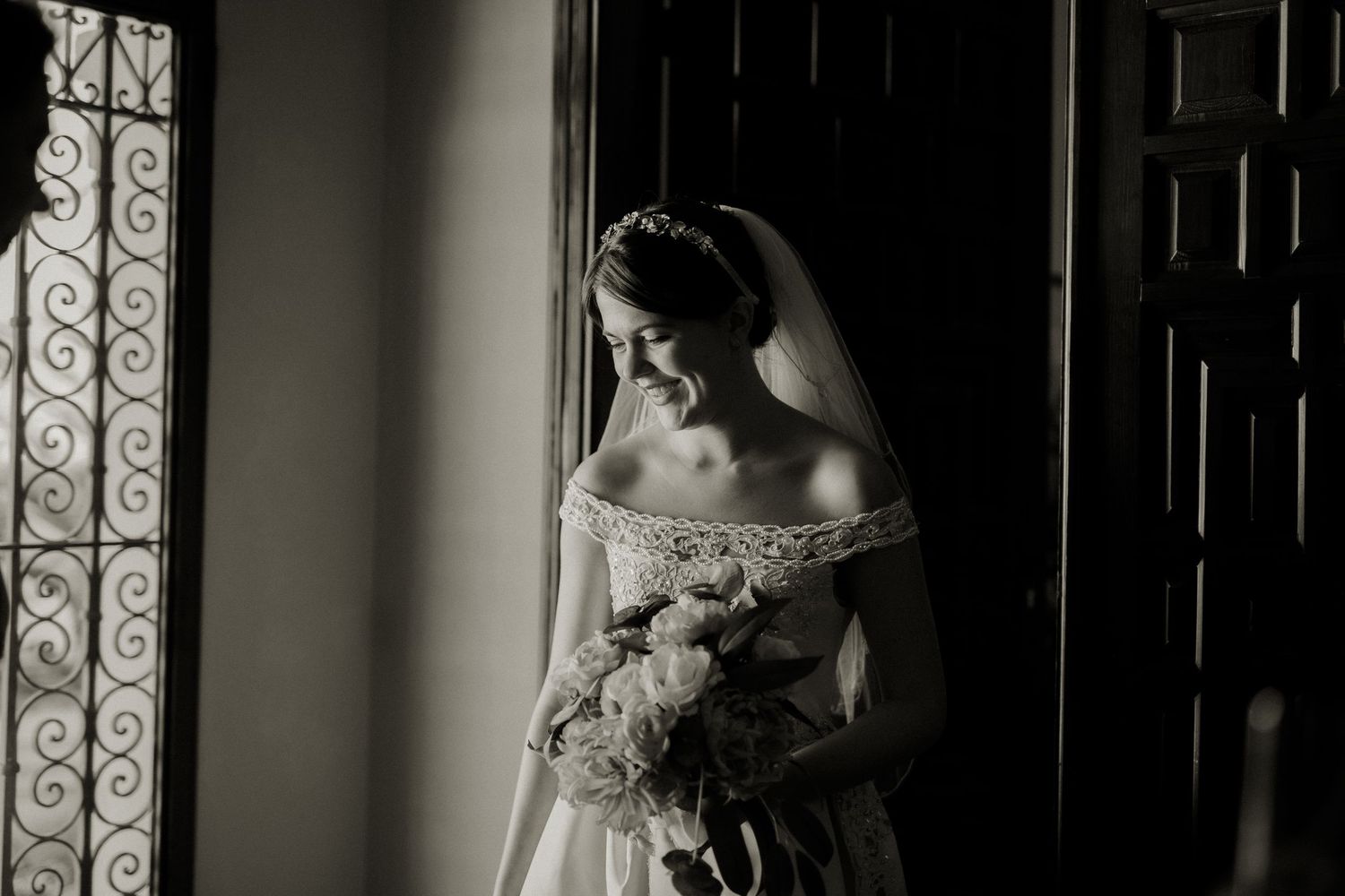 Bride in off-shoulder lace wedding dress holding bouquet, standing by window in black and white photo.