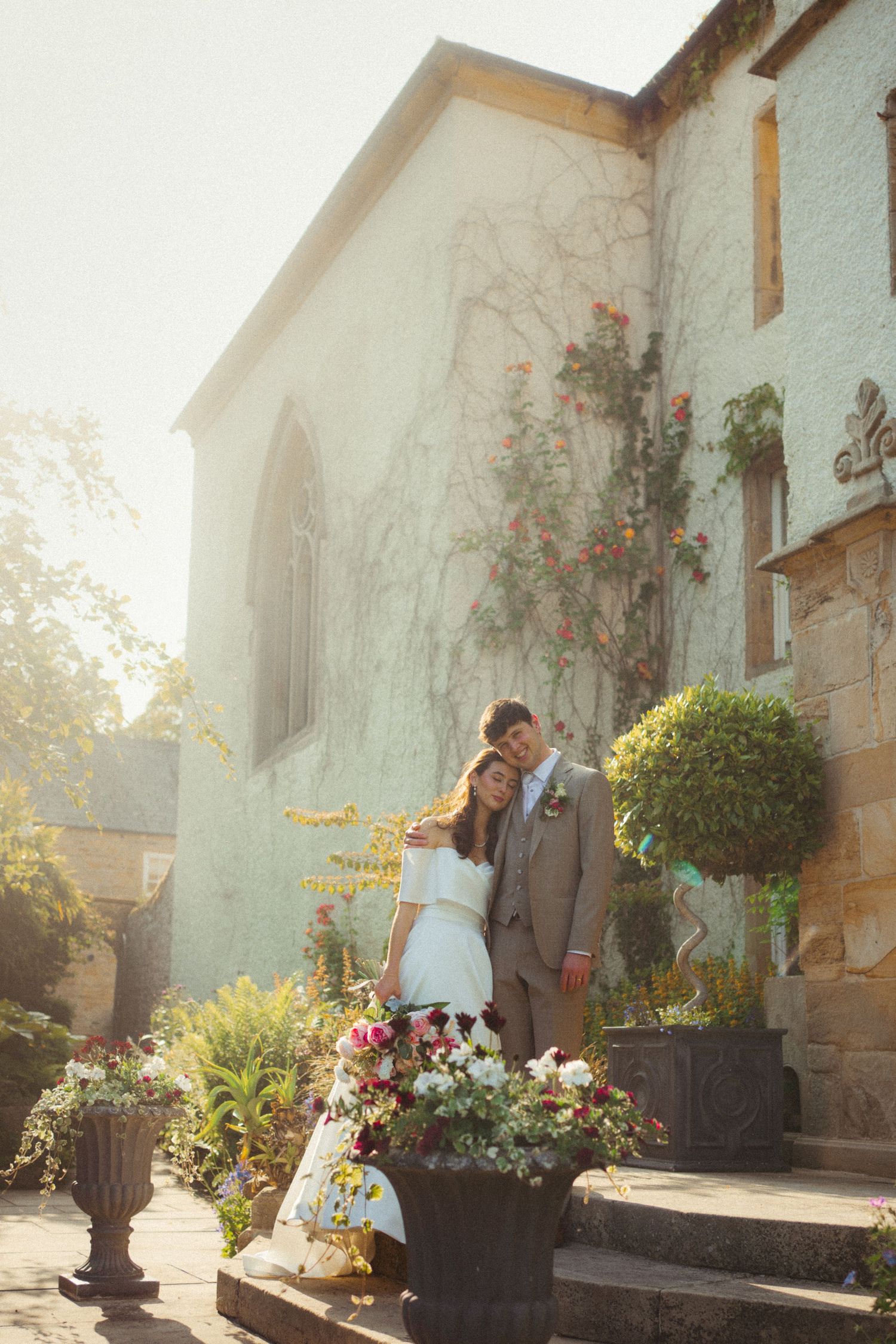 Bride and groom embracing on sunlit steps outside ivy-covered stone manor wedding venue.