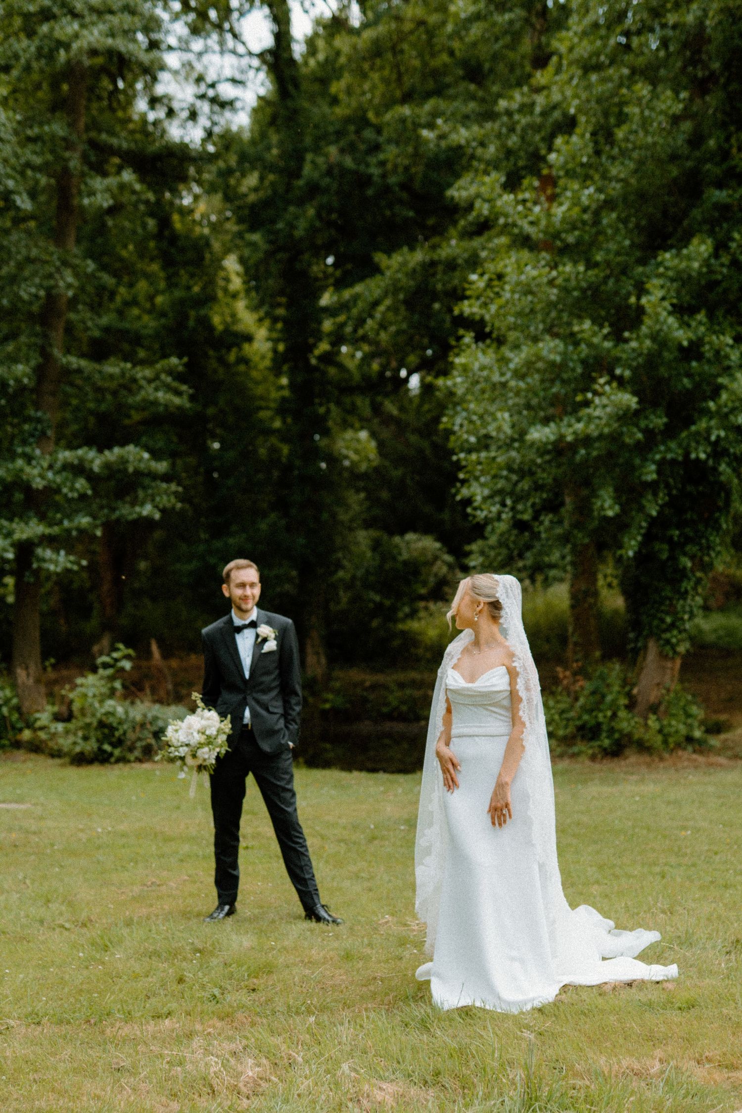 Bride and groom standing apart in a green garden, bride wearing a white cape wedding gown with bouquet.