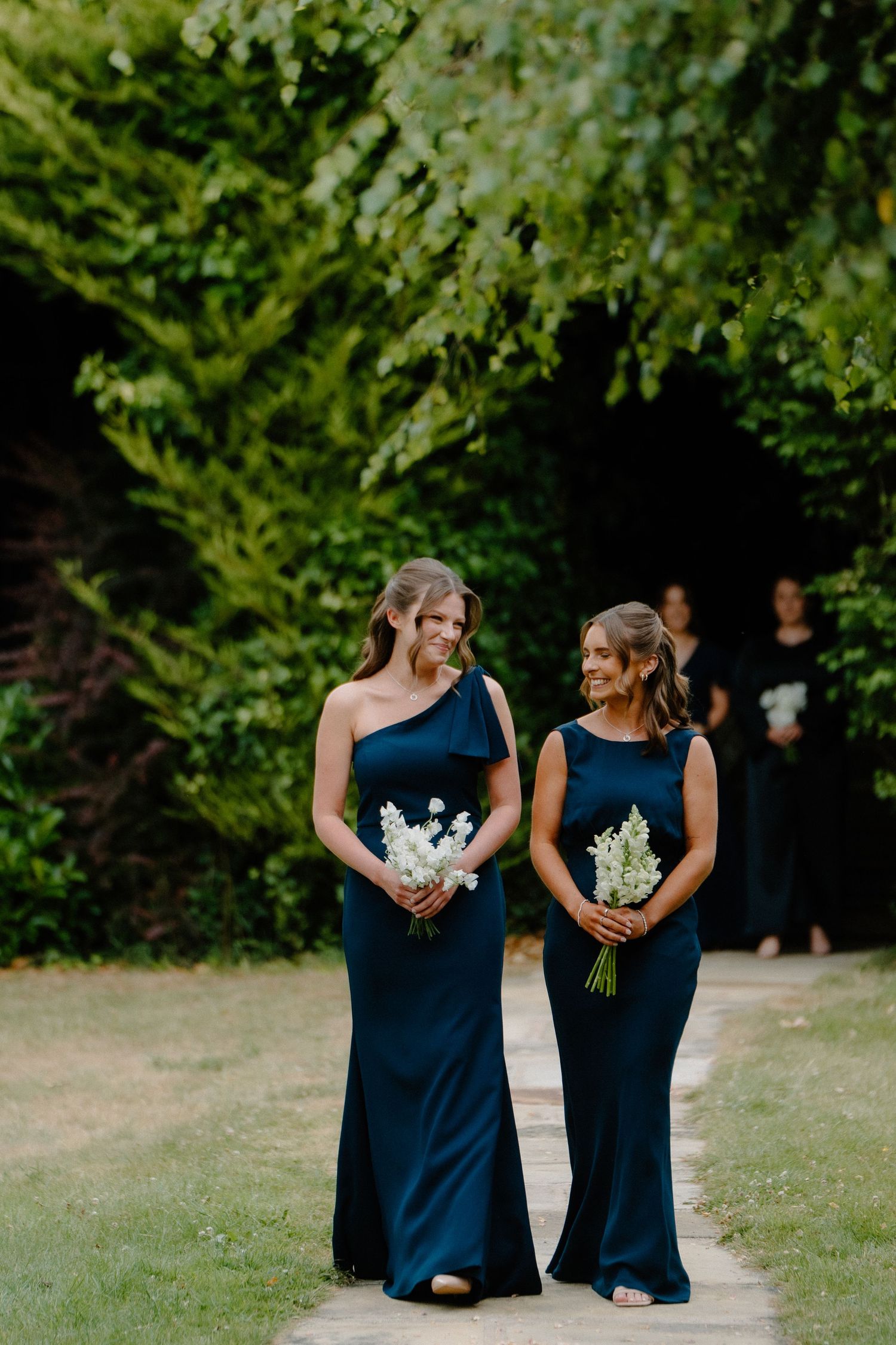 Two bridesmaids in navy blue gowns carry white floral bouquets while walking along garden pathway.