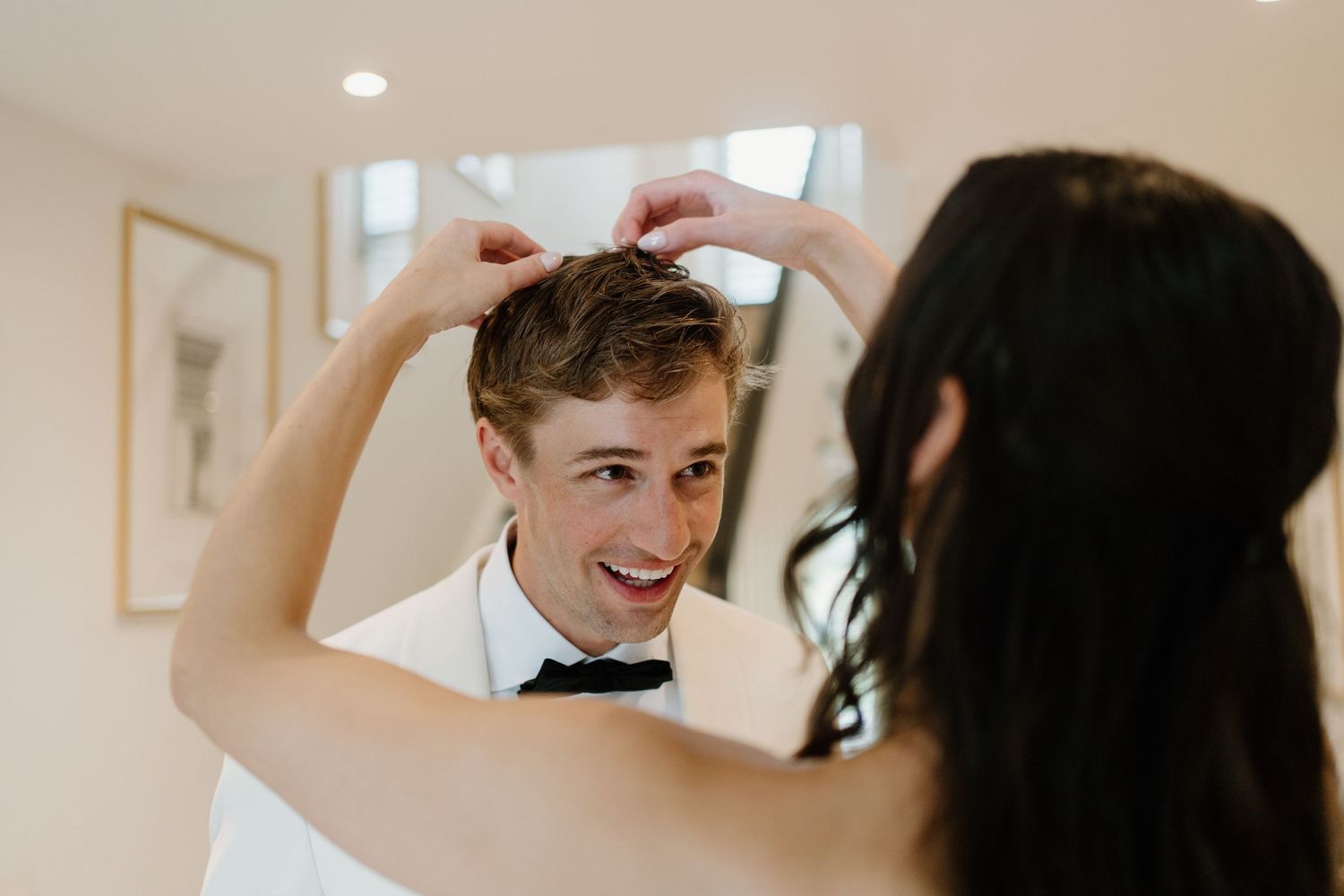 Groom in white tuxedo jacket smiling while bride fixes his hair before wedding ceremony.