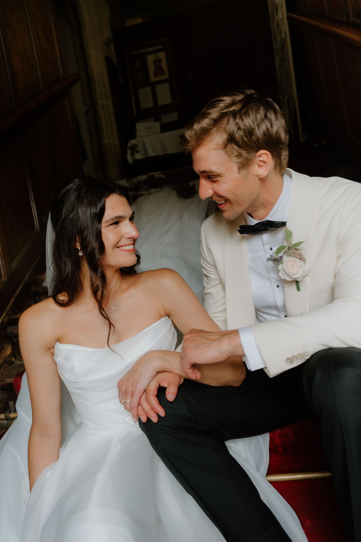 Bride and groom sit closely together in a warmly lit rustic interior, smiling and holding hands tenderly.