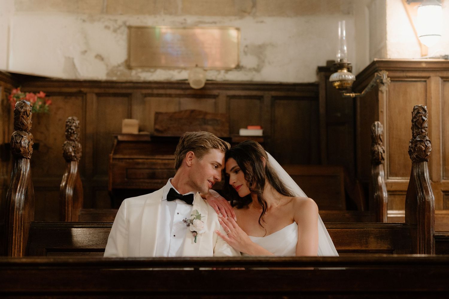 Bride and groom in white tuxedo sharing intimate moment in rustic wooden church pew.