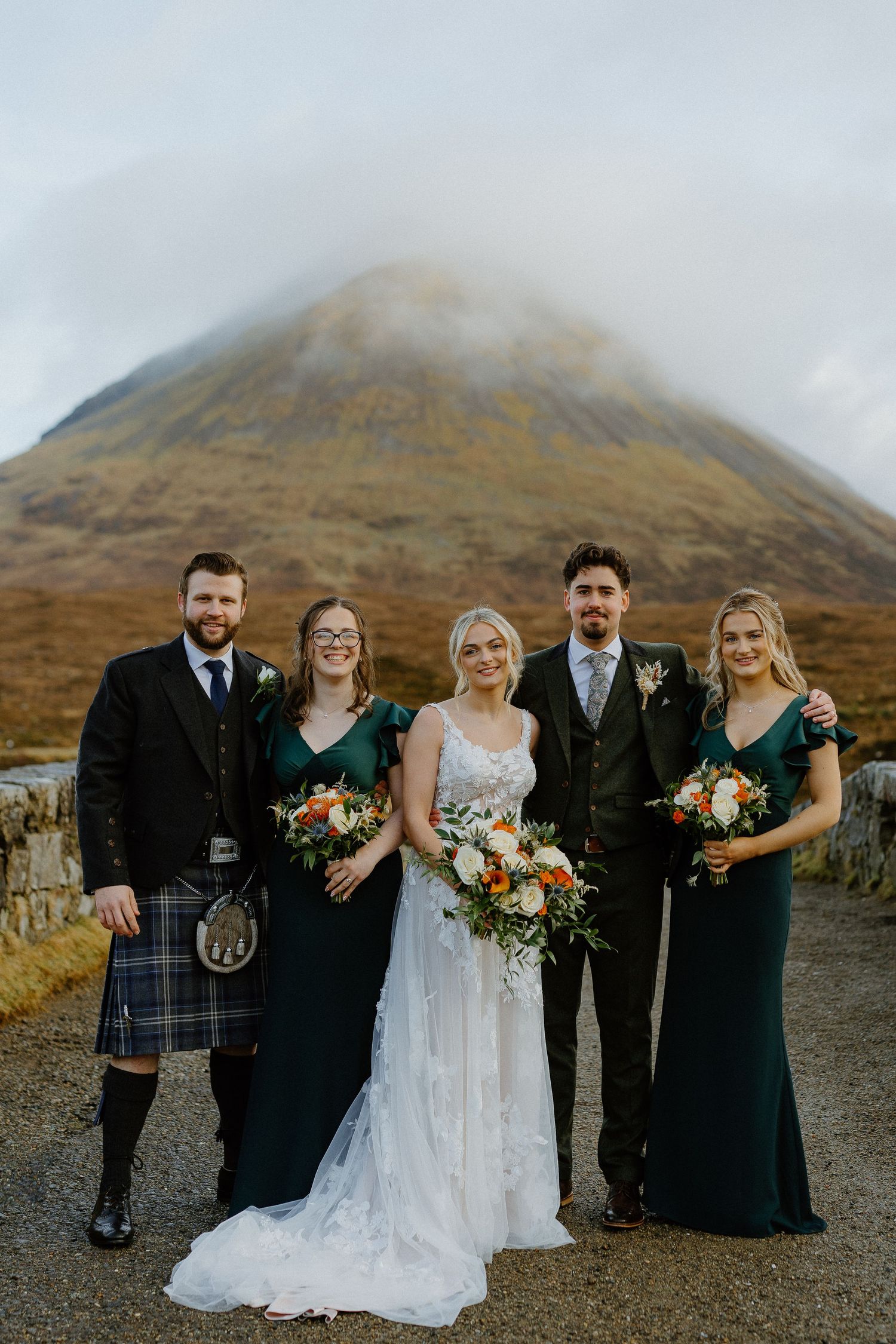 Wedding party poses before misty Scottish Highland mountain, bride in lace gown with groomsmen in kilts and bridesmaids in emerald dresses.
