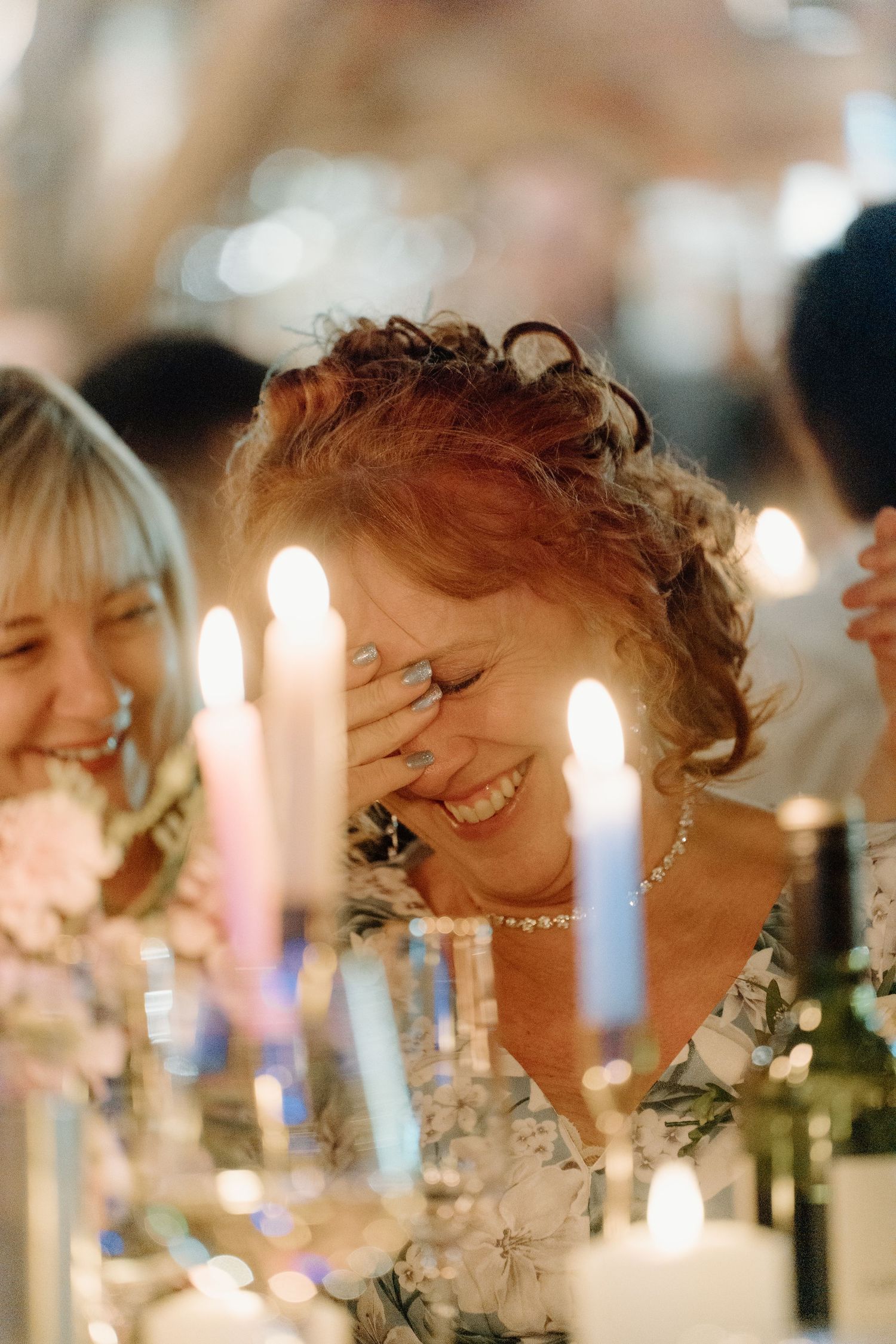 A joyful wedding guest laughs warmly while seated at a candlelit reception table with floral arrangements.