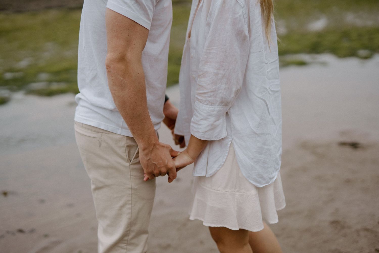 Close-up of a couple holding hands on a sandy beach, both wearing white outfits.