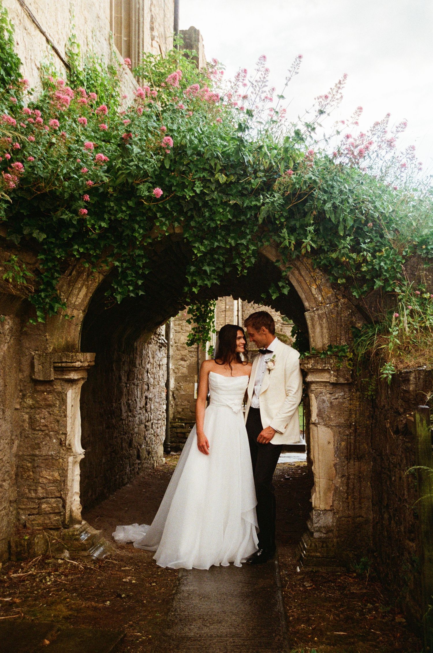 Bride and groom embrace under a stunning rose-covered stone archway at a historic wedding venue.