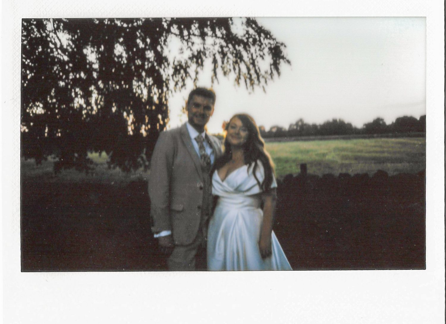 Bride and groom pose together under weeping willow tree in vintage Polaroid style photo at outdoor wedding.