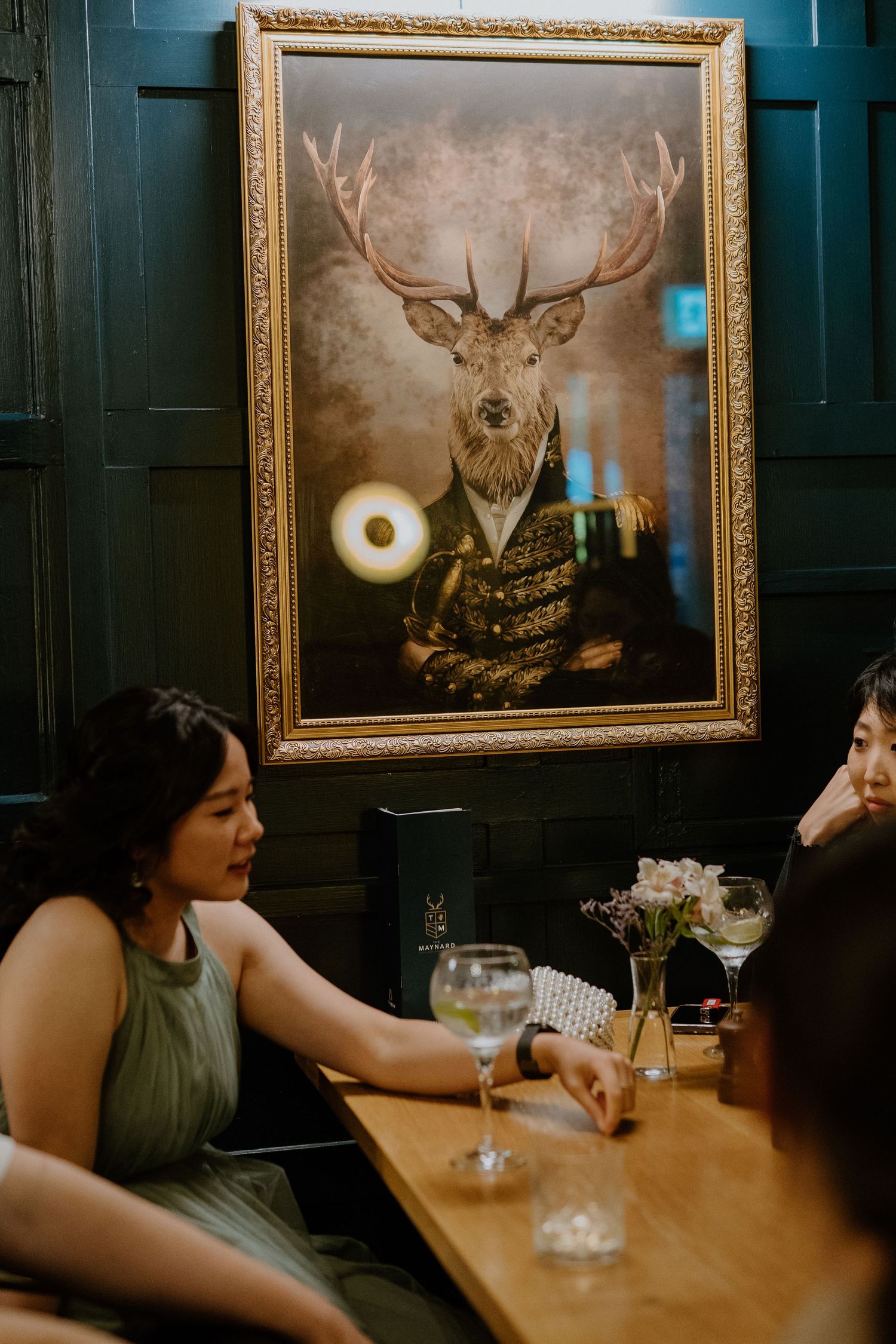 Wedding guests socializing at dark wood bar beneath ornate stag portrait painting.