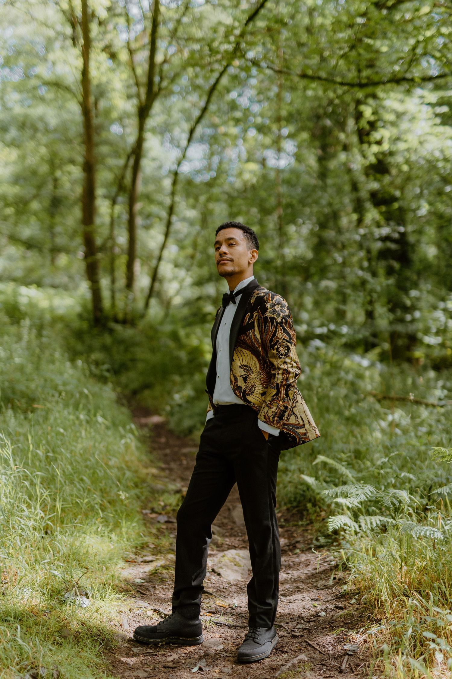 Groom in ornate gold batik blazer stands on forest path surrounded by lush green trees.