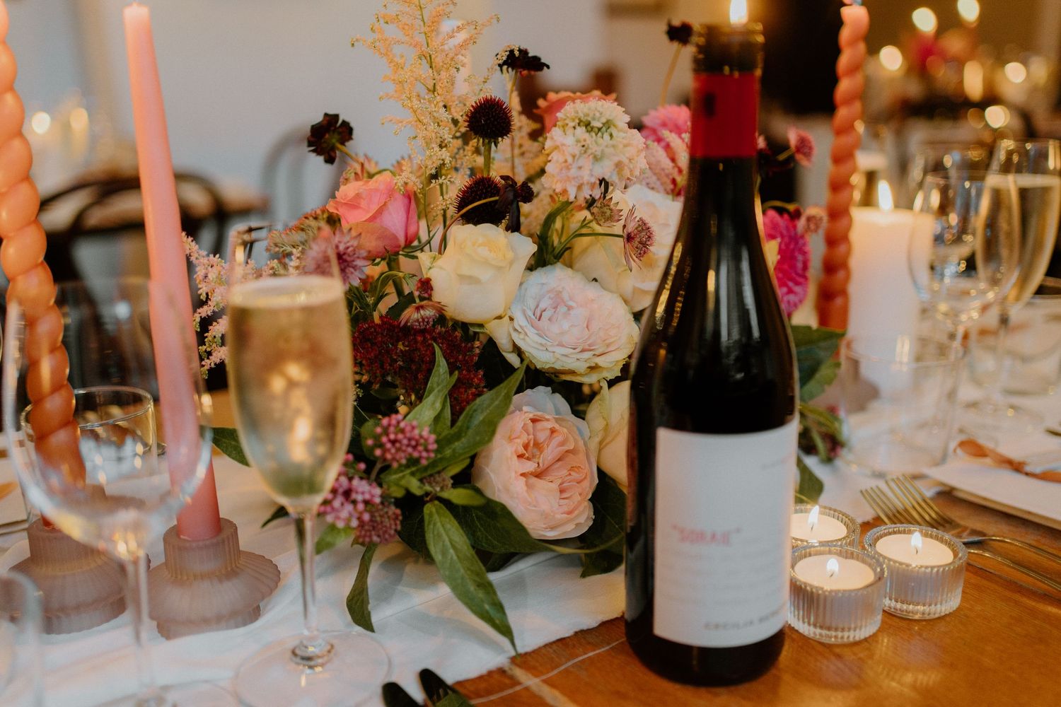 Elegant wedding table setting with pink floral centerpiece, candles, champagne flutes, and red wine bottle.