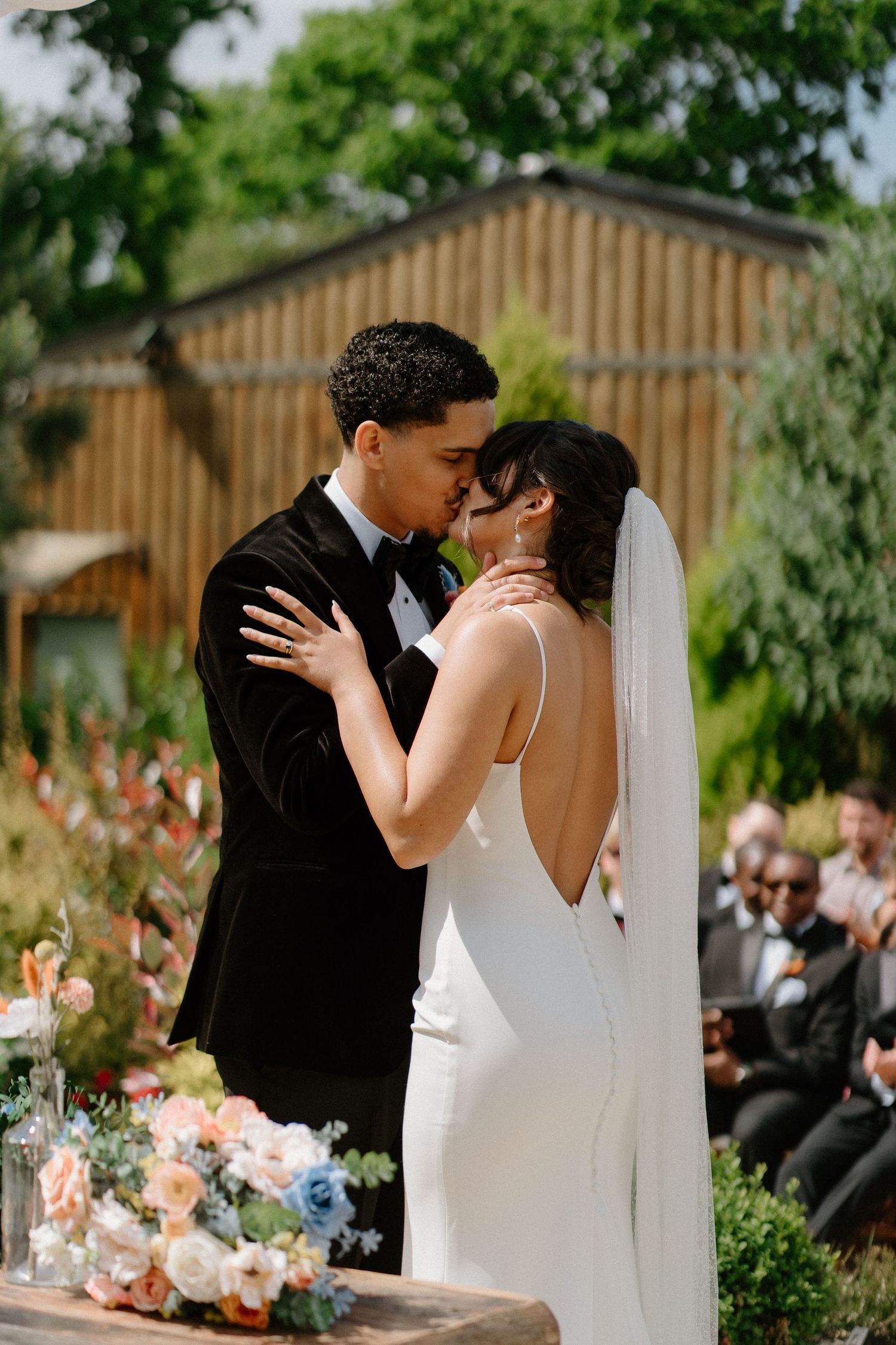 Bride and groom sharing their first kiss at outdoor wedding ceremony surrounded by colorful floral arrangements.