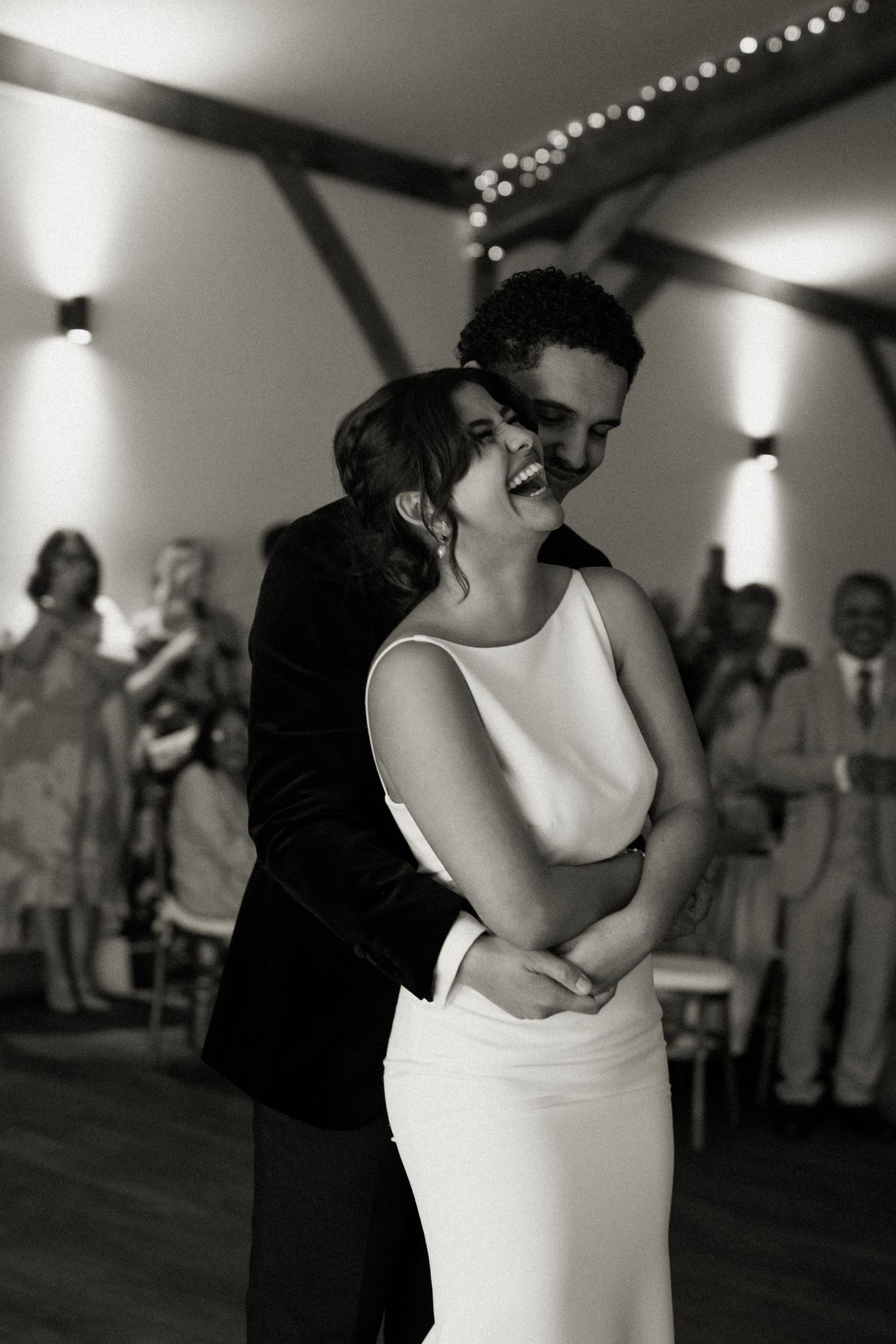 Black and white photo of groom embracing laughing bride during first dance while guests celebrate in background.