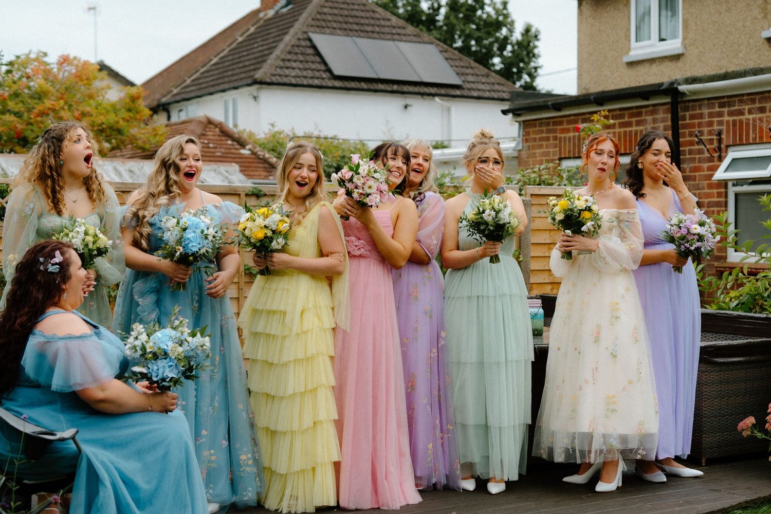 Bridesmaids in pastel dresses of blue, yellow, pink, mint and lavender holding floral bouquets stand together outdoors.