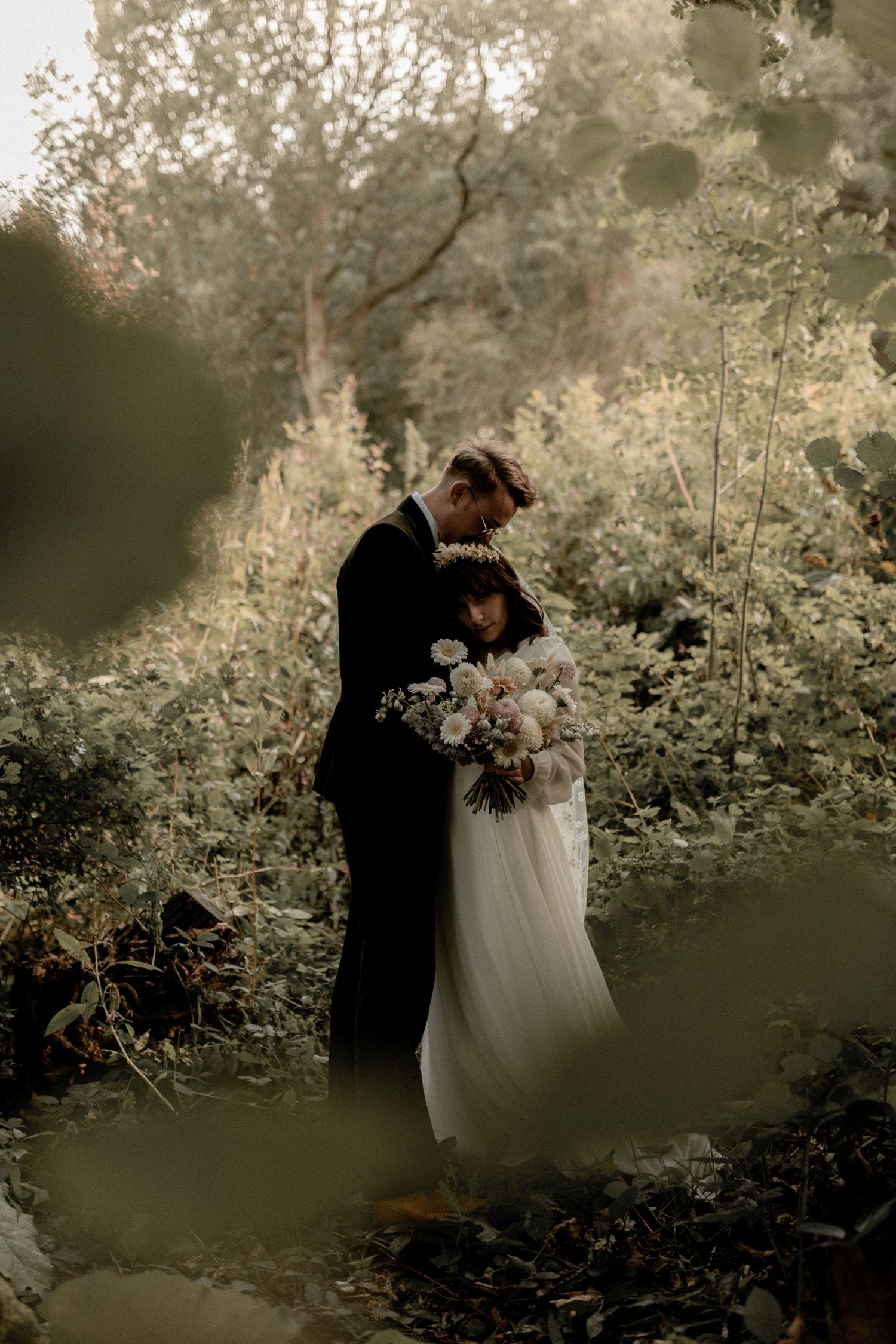 Moody wedding portrait of a couple in formal attire surrounded by lush greenery.