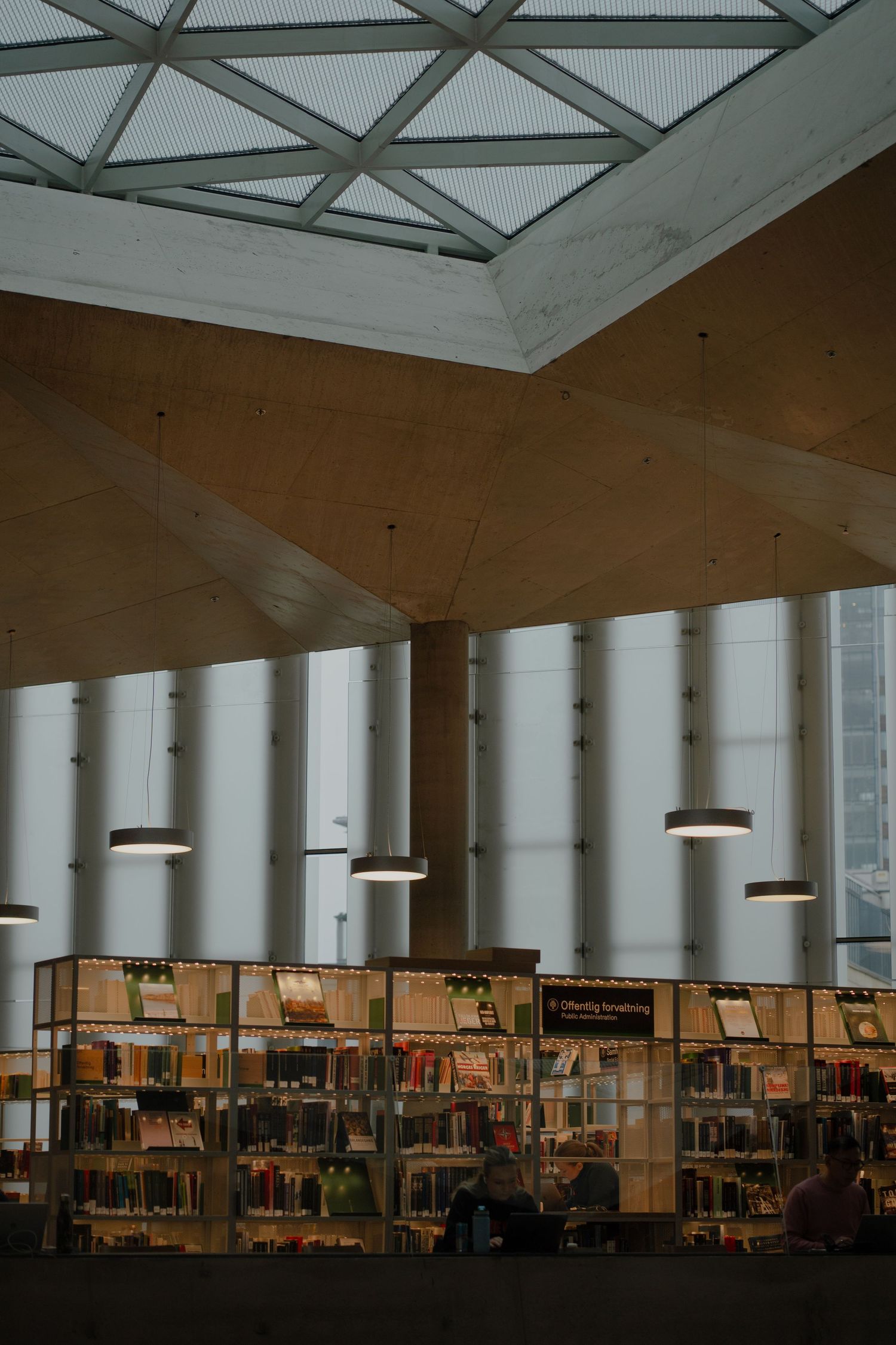 Modern library interior with hanging circular lights and wall-to-wall bookshelves under a geometric skylight ceiling.