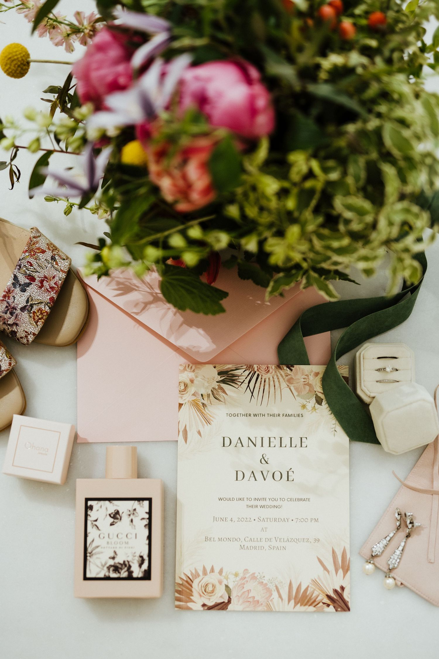 Elegant bohemian wedding invitation for Danielle and Davoe surrounded by pink peonies and Gucci perfume flatlay.