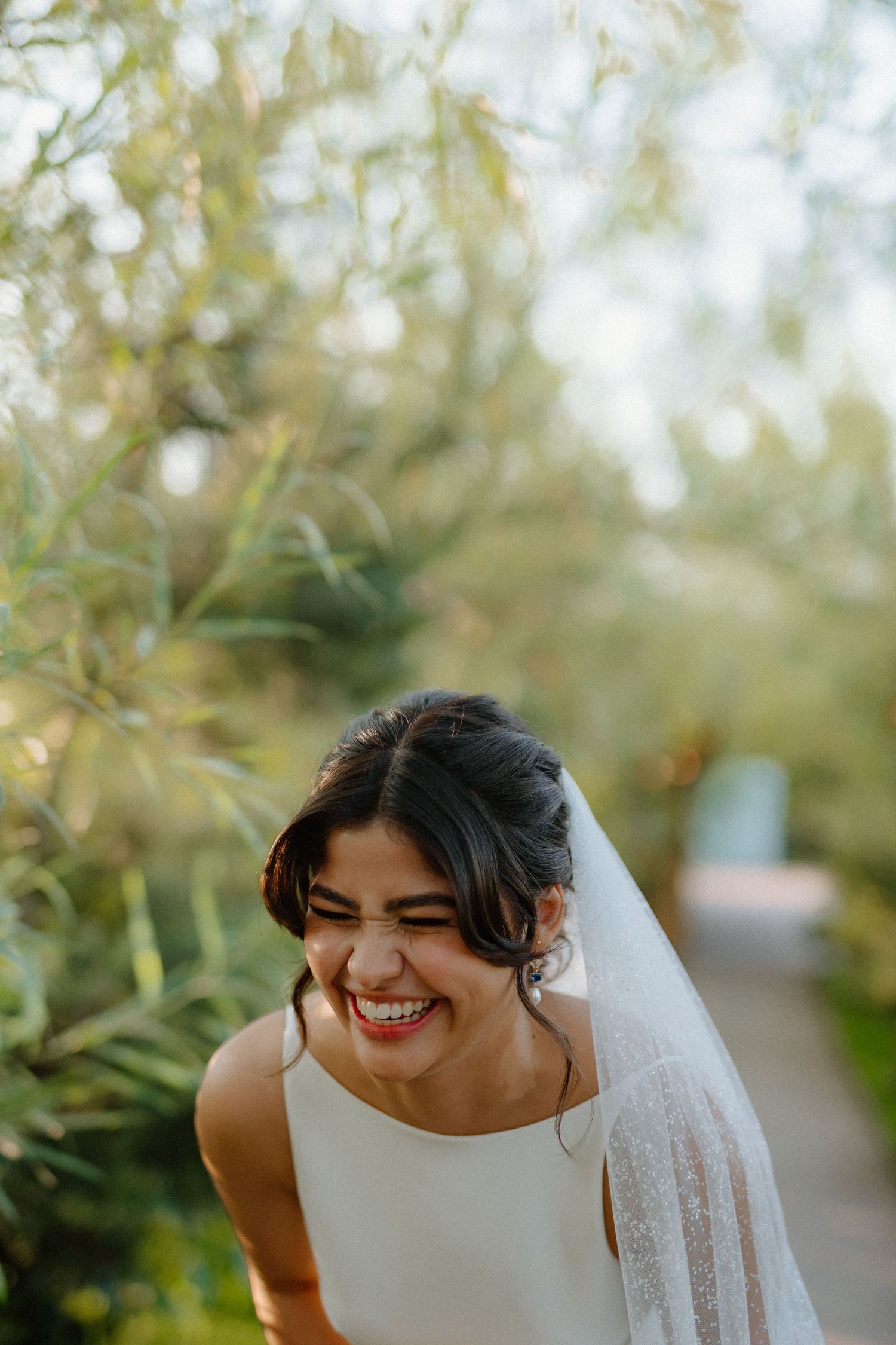 Radiant bride in white dress laughing joyfully outdoors with veil and soft natural light.