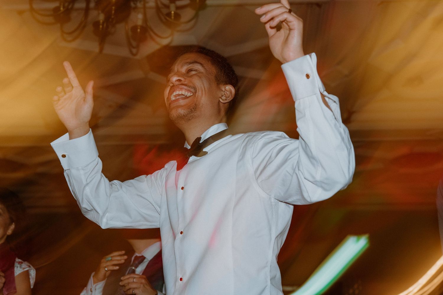 Groom in white dress shirt dancing enthusiastically with arms raised at wedding reception party.