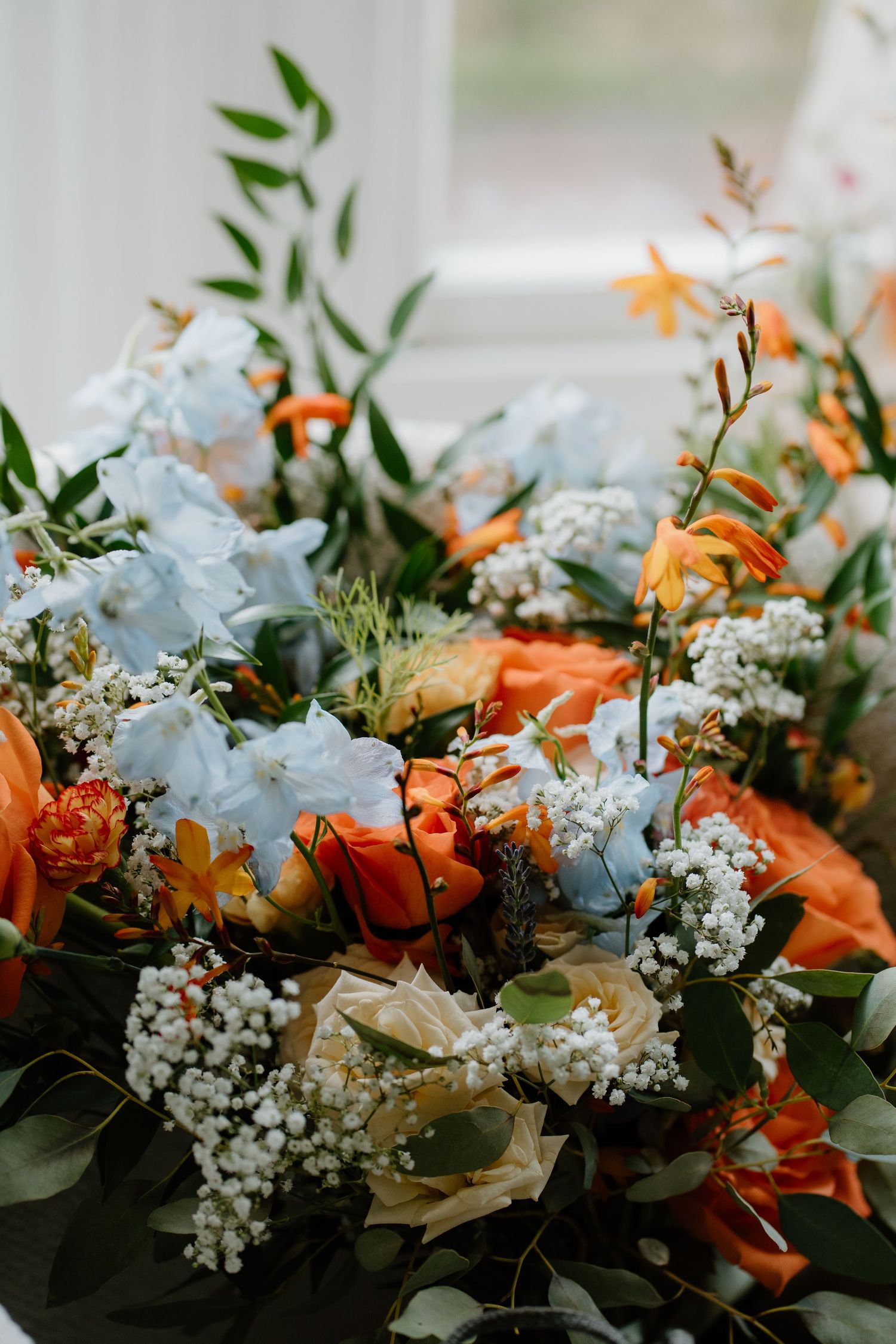 Lush wedding bouquet featuring orange roses, blue hydrangeas, white gypsophila, and green foliage.