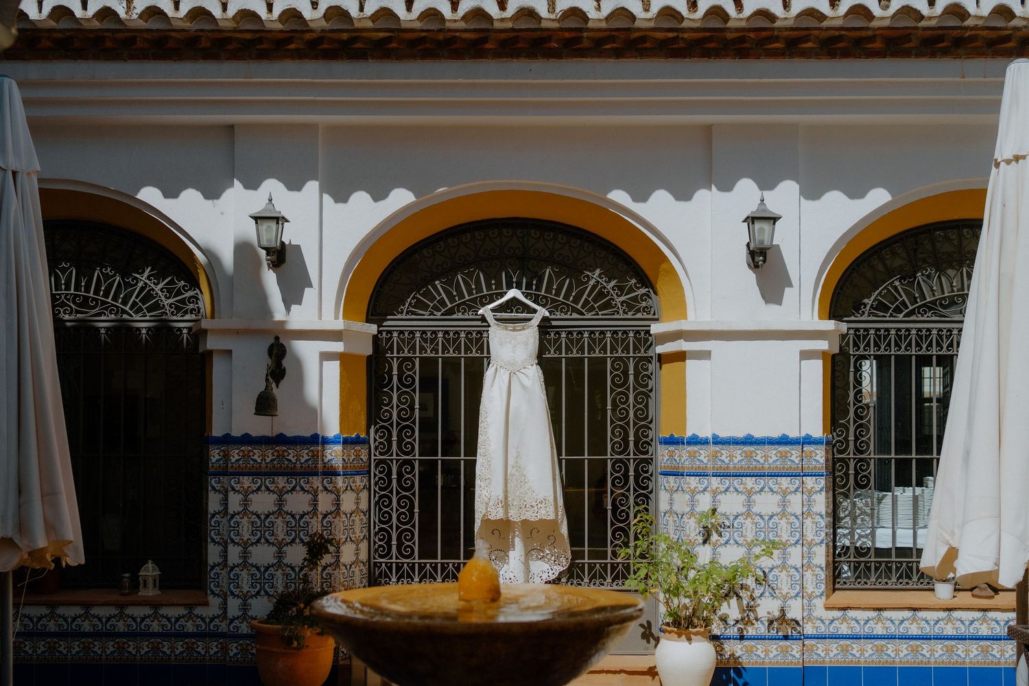 Elegant white wedding dress hangs in Spanish courtyard with ornate tiled walls and fountain.