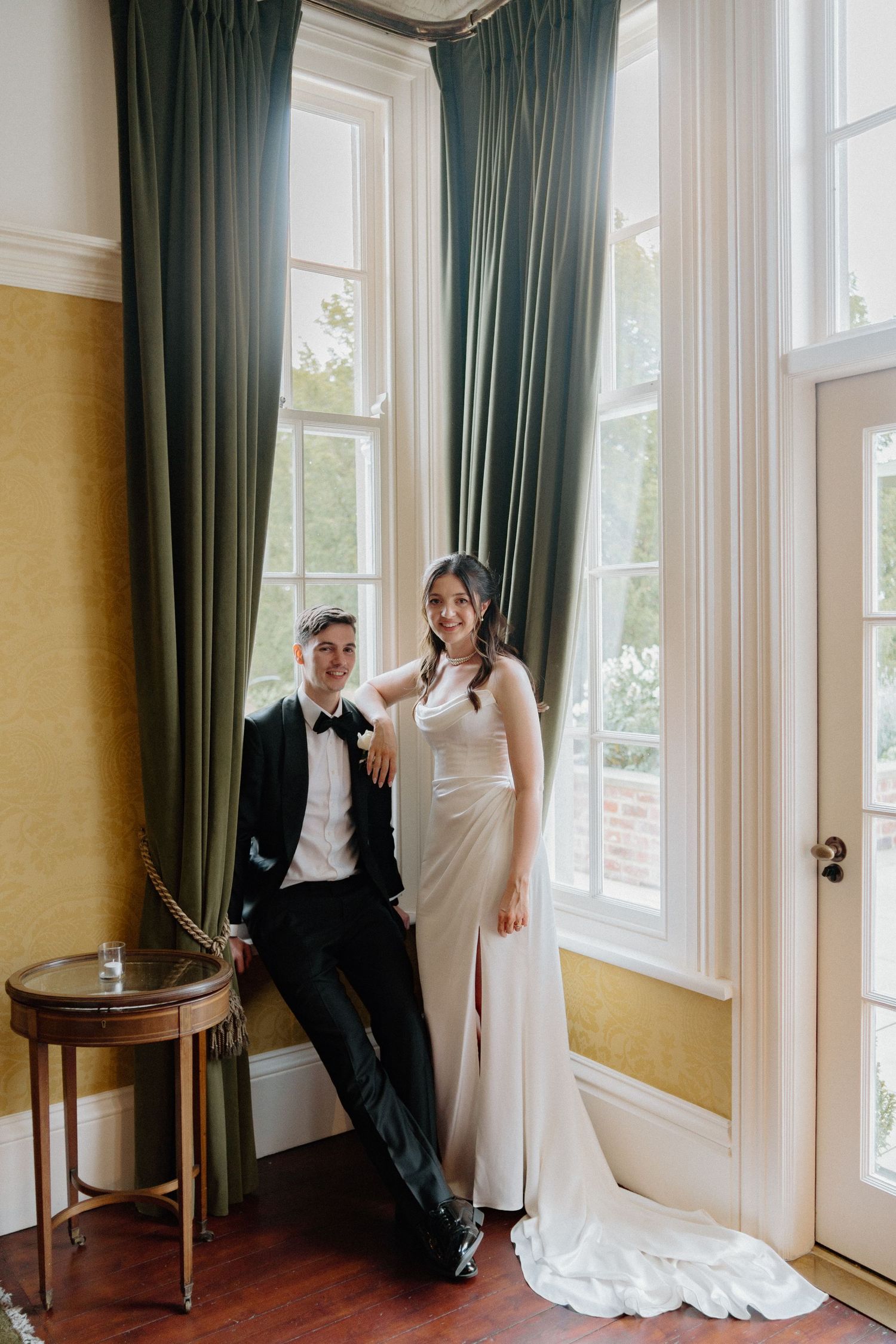 Elegant bride in sleek white gown and groom in black tuxedo posing by tall windows with green drapes.
