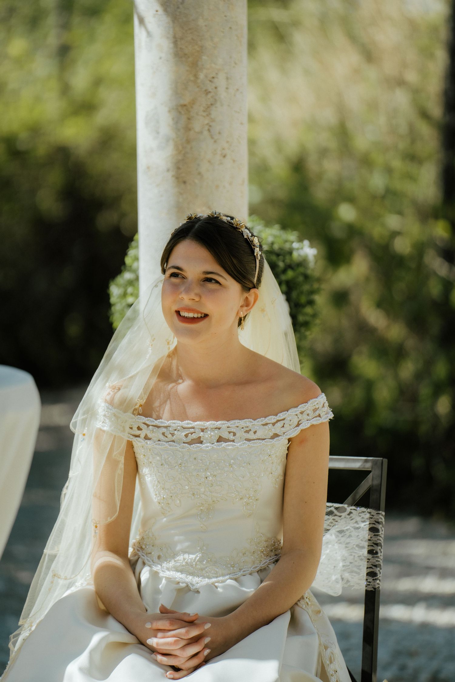 Smiling bride in an off-shoulder lace wedding gown with veil sits during an outdoor garden ceremony.