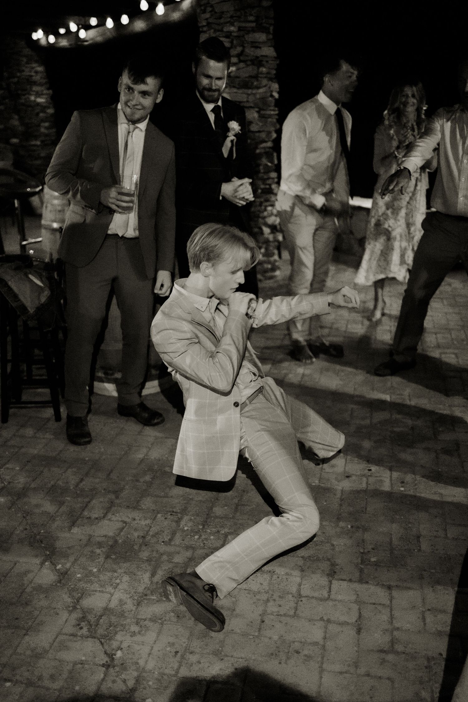 A young boy in a suit energetically dancing at a wedding reception while guests watch and celebrate in the background.