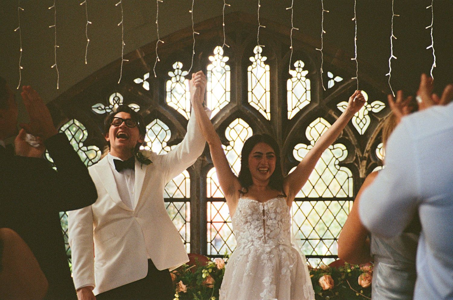 Bride and groom celebrating joyfully before a stained glass church window with raised hands.